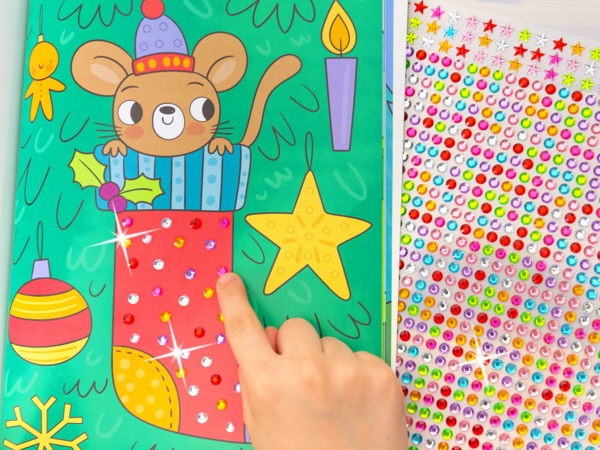 Es wird eine Innenseite des Buches „Mein Diamant-Stickerheft – Wintertraum“ in Aktion gezeigt: Eine Kinderhand ragt von unten in das horizontale Bild rein und klebt soeben einen Diamantsticker auf die Innenseite. Auf dem Bild sind bereits alle Felder mit Diamanten geschmückt und glänzt daher wunderschön. Unterhalb des Heftes ragt der Stickerbogen hervor und zeigt eine große Vielzahl an bunten Diamantstickern.