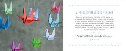 Gezeigt wird eine Innen- oder Zusatzansicht von „Gute Besserung – Alles Liebe!“. Auf der linken Seite hängen bunt gefaltete Papierkraniche in verschiedenen Farben wie Rot, Blau, Grün und Pink vor einem grauen Hintergrund. Die rechte Seite ziert ein eleganter Text in Türkis mit der Überschrift „Verleih deinem Glück Flügel“ sowie einem Zitat von J. M. Barrie. Die Schrift ist klar und modern, um eine freundliche und optimistische Stimmung zu vermitteln.
