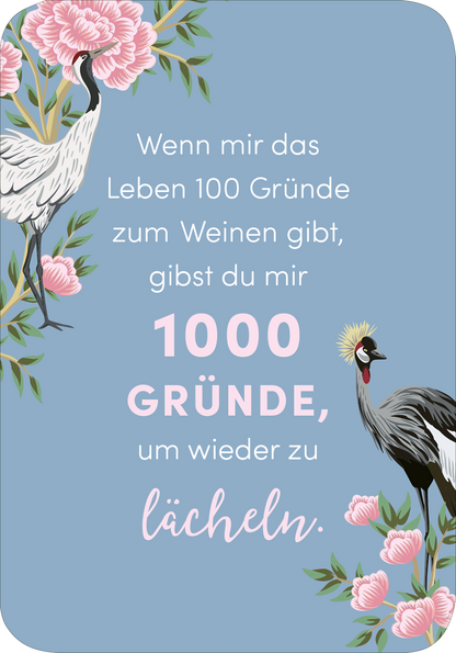 Gezeigt wird eine Innen- oder Zusatzansicht von „Weil du eine wundervolle Frau bist!“. Der Hintergrund ist in einem sanften Blau gehalten und mit rosa Pfingstrosen und stilisierten Kranichen verziert. Der Text ist in unterschiedlichen Schriftgrößen und -arten gestaltet: „Wenn mir das Leben 100 Gründe zum Weinen gibt,“ in einer eleganten, serifenlosen Schrift, und „1000 GRÜNDE, um wieder zu lächeln“ in einer größeren, verspielten Schriftart. Das Gesamtbild strahlt eine...