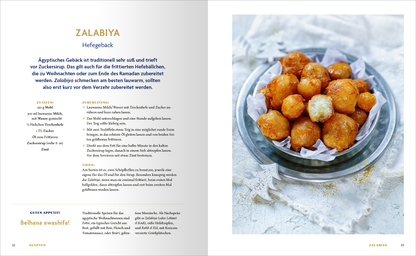 Gezeigt wird eine Innen- oder Zusatzansicht von „Weihnachten rund um die Welt“. Auf der linken Seite sind die Rezeptdetails für „Zalabiya“ abgebildet, übersichtlich in dunkler Schrift auf hellem Hintergrund, mit goldenen Akzenten im Titel. Auf der rechten Seite ist ein transparentes Glas mit rundem, golden-braunem Gebäck gefüllt, dekorativ angerichtet auf einer platzierten Serviette. Im Hintergrund ist eine helle, schlicht gehaltene Tischoberfläche zu sehen, die die Leckerei...