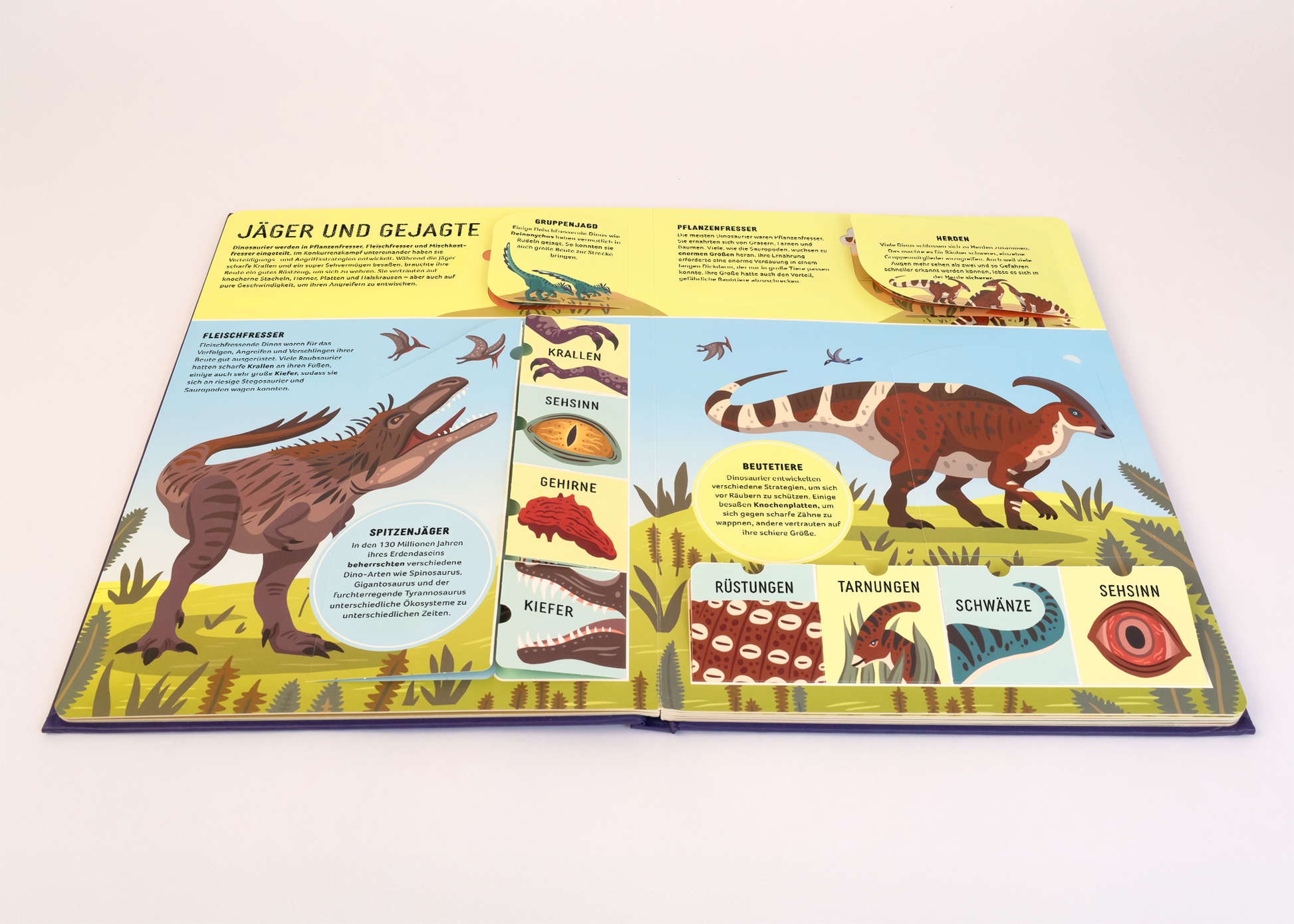 Gezeigt wird eine Innenansicht von „Dinosaurier“. Die Doppelseite präsentiert eine farbenfrohe Illustration mit verschiedenen Dinosauriern in lebendigen Grüntönen und Erdfarben. Der obere Bereich zeigt die Überschrift „Jäger und Gejagte“ in auffälliger, verspielter Typografie. Links sind erläuternde Kästen zu einer Vielzahl von Dinosaurierarten, ihre Merkmale und Lebensweisen auf hellem Hintergrund angeordnet. Die bildhaften Darstellungen sind klar und ansprechend gestaltet, um...