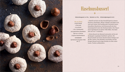 Gezeigt wird eine Innen- oder Zusatzansicht von „Vanille, Zimt und Weihnachtsduft“. Auf der linken Seite sind mehrere runde, helle Haselnussplätzchen zu sehen, die jeweils mit einer ganzen Haselnuss darauf dekoriert sind. Der Hintergrund ist dunkel und strukturiert, was einen ansprechenden Kontrast zu den Plätzchen bildet. Rechts befindet sich ein Rezepttext in eleganter, goldfarbener Schrift, der auf einem sanft rosa Hintergrund steht und eine leicht lesbare, moderne Typografie verwendet.