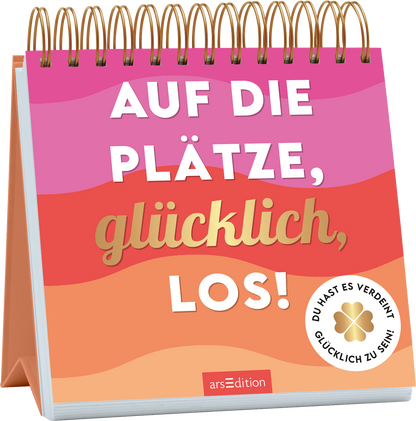 Das Buchcover zeigt eine spiralförmige Bindung mit leuchtend bunten Farbverläufen in Pink- und Orangetönen, die eine fröhliche Atmosphäre schaffen. In der Mitte prangt der Titel „AUF DIE PLÄTZE, glücklich, LOS!“ in einer großen, weißen und goldenen Schrift. Auf der unteren rechten Seite befindet sich ein weißes Label mit der Aufschrift „DU HAST ES VERDIENT, GLÜCKLICH ZU SEIN!“. Ganz unten mittig steht der Logoschriftzug von arsEdition.