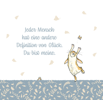 Gezeigt wird eine Innen- oder Zusatzansicht von „Weißt du eigentlich, wie lieb ich dich hab? 365 Liebesbotschaften für dich“. Die Illustration zeigt einen fröhlichen Hasen, der mit outgestreckten Pfoten springt, um eine Schmetterling zu fangen. Der Hintergrund ist hell und sanft in Weiß gehalten, während der untere Teil in einem zarten Blau mit floralen Mustern gestaltet ist. Der Text in geschwungener, grauer Schrift hebt die Botschaft hervor: „Jeder Mensch hat eine andere Definition...