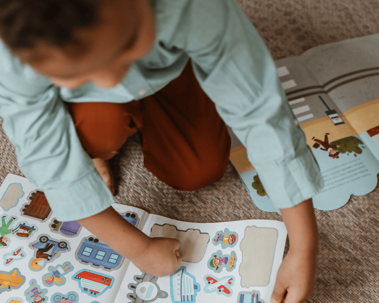 Ein Kind sitzt auf einem Teppich und beschäftigt sich mit einem Stickerbuch, in dem bunte Sticker mit Motiven wie Fahrzeugen, Figuren und Objekten zu sehen sind. Auf der Seite daneben liegt ein aufgeschlagenes Pappbuch mit einer illustrierten Szene, die eine Straße und Bäume zeigt. Das Kind trägt ein langärmliges, mintfarbenes Hemd und eine rote Hose.
