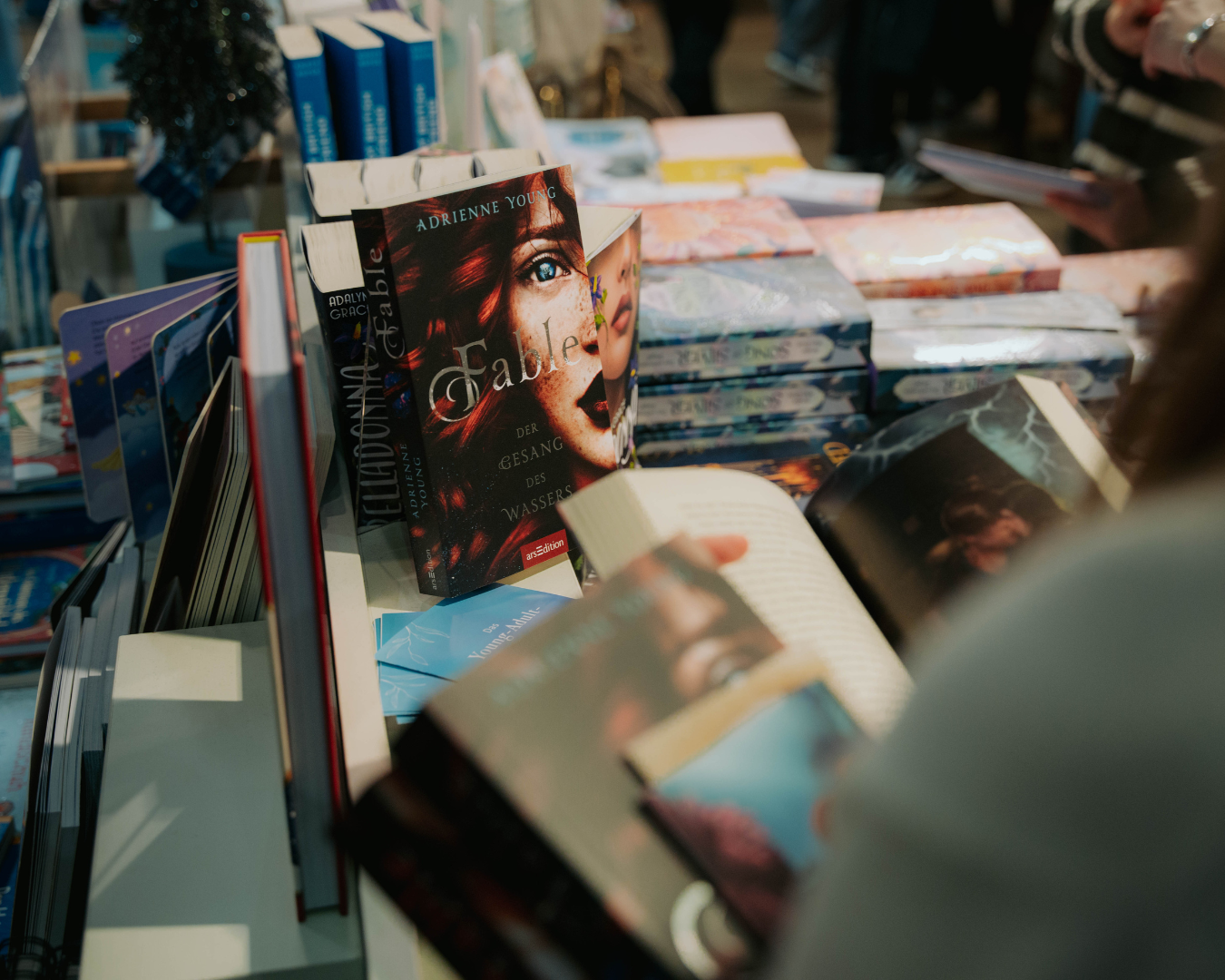 Aufnahme eines Büchertisches auf einer Buchmesse oder in einem Buchladen. Im Vordergrund sind mehrere aufgeschlagene Bücher zu sehen, die von einer Person durchgeblättert werden. Im Zentrum steht das Buch ‚Belladonna – Wassers der Finsternis‘ von Adalyn Grace, aufrecht in einem Stapel präsentiert. Im Hintergrund befinden sich weitere gestapelte Bücher und ein verschwommener Bereich mit weiteren Besuchern.