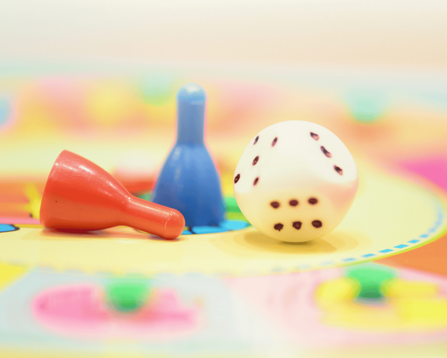 Das Bild zeigt ein buntes Brettspiel-Spielfeld mit einem weißen Spielwürfel und zwei Spielfiguren in den Farben Rot und Blau. Die rote Spielfigur liegt umgekippt, während die blaue aufrecht steht. Der Hintergrund ist unscharf und zeigt helle, farbenfrohe Muster, die eine fröhliche Atmosphäre vermitteln.