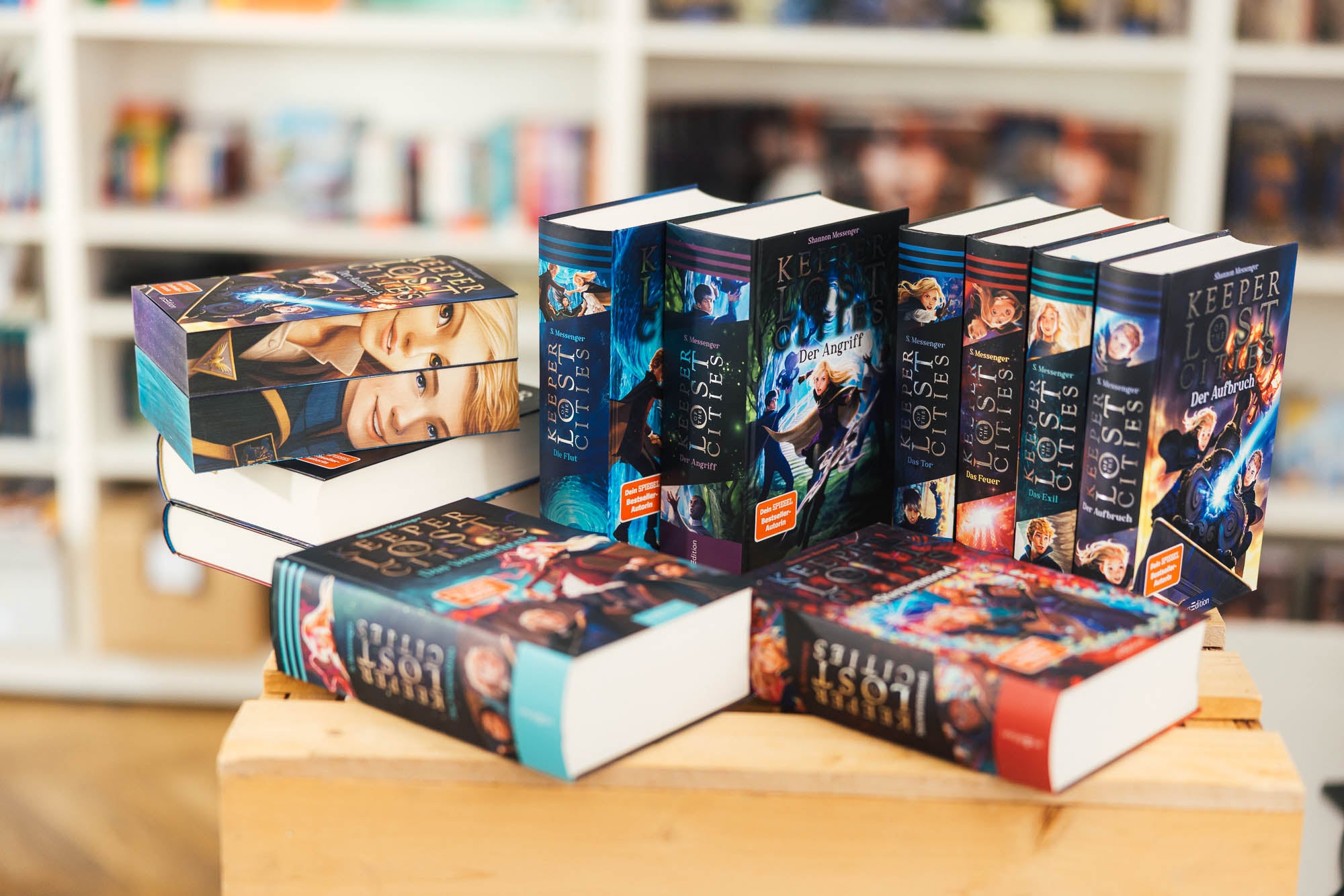 Ein Stapel gebundener Bücher der Buchreihe „Keeper of the Lost Cities“ von Shannon Messenger auf einem Holztisch in einem Bücherladen. Die Bände sind farbenfroh gestaltet, mit Illustrationen und Titeln wie „Der Aufbruch“ und „Der Angriff“. Im Hintergrund unscharf zu erkennen sind Bücherregale mit weiteren Büchern.