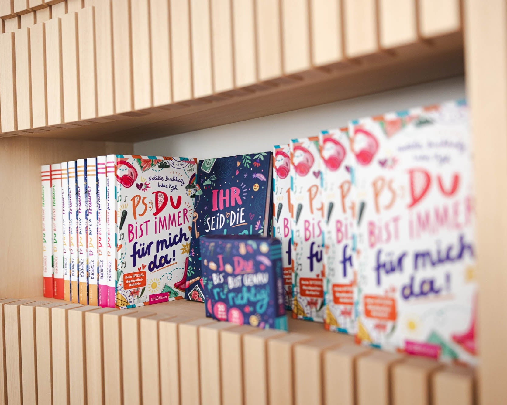 Ein Regal aus hellem Holz mit mehreren Büchern, die in zwei Reihen aufgestellt sind. Links stehen Bücher mit bunten Rücken in einer Reihe, rechts sind mehrere Exemplare des Buches „PS: Du bist immer für mich da!“ mit farbenfrohem Cover zu sehen. Dazwischen befindet sich ein kleineres Buch mit dem Titel „Ihr seid die Besten“. Die Gestaltung der Buchcover ist verspielt und lebendig.