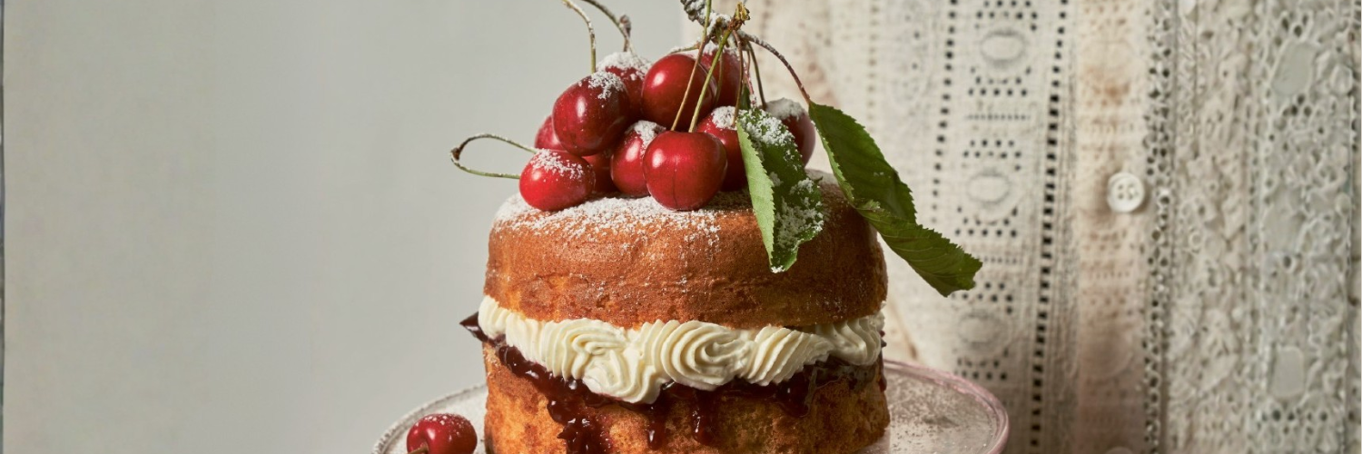 Alt-Text: Ein zweischichtiger Kuchen, gefüllt mit Sahne und Kirschmarmelade, steht auf einer rosa Glasplatte. Der Kuchen ist mit frischen roten Kirschen, grünen Blättern und einer leichten Schicht Puderzucker dekoriert. Im Hintergrund ist ein Teil eines weißen, bestickten Kleidungsstücks sichtbar, das eine elegante und rustikale Atmosphäre vermittelt.