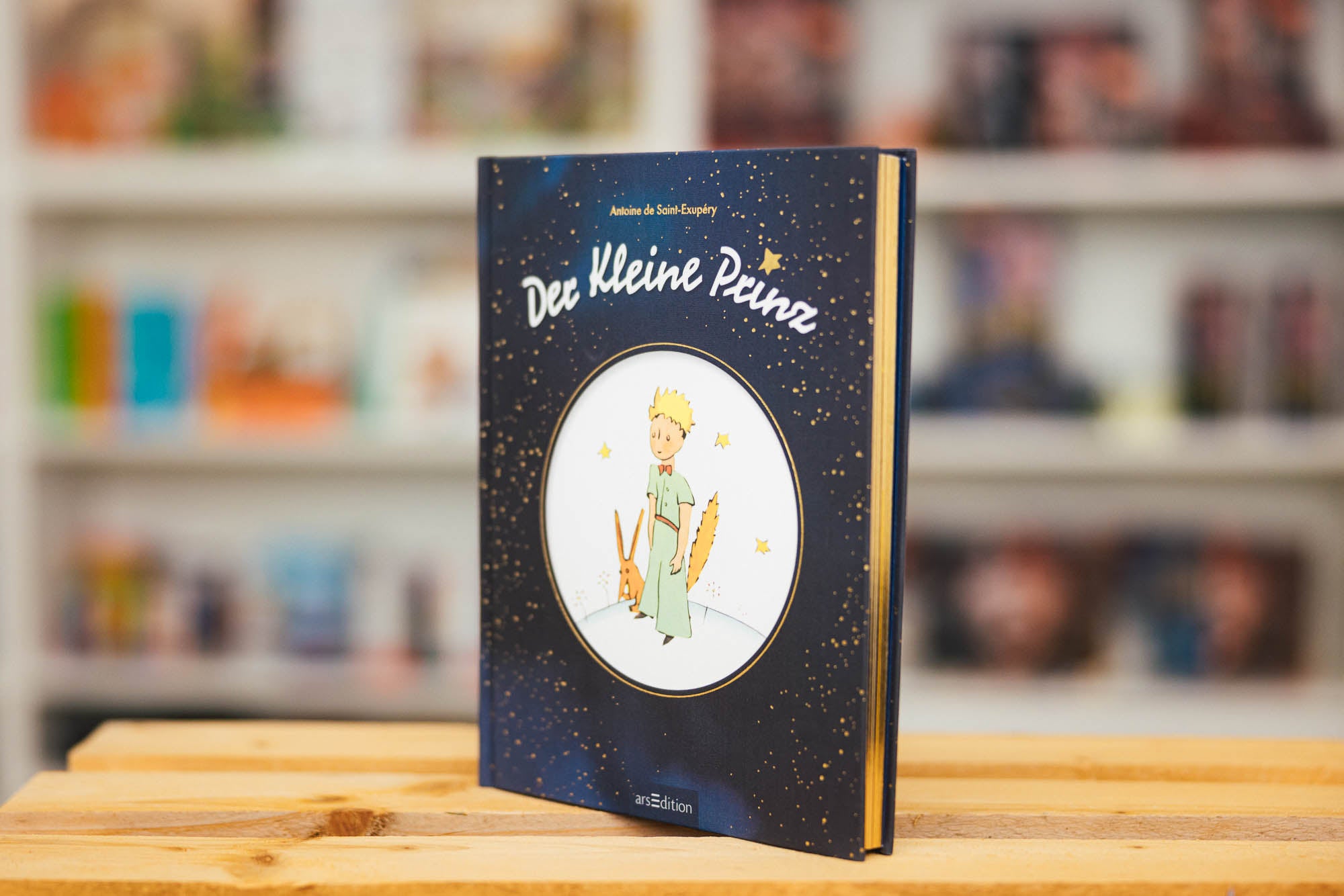 Eine gebundene Ausgabe des Buches „Der kleine Prinz“ von Antoine de Saint-Exupéry, ausgestellt auf einem Holztisch. Das Buchcover ist dunkelblau mit goldenen Sternen und zeigt eine Illustration des kleinen Prinzen mit einem Fuchs. Im Hintergrund sind unscharf Bücherregale mit weiteren Büchern zu erkennen.