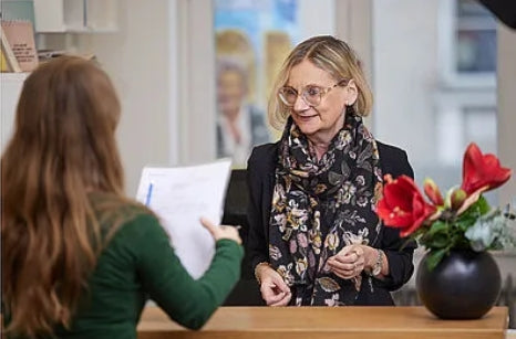 Eine Frau in grüner Kleidung steht an einem Empfangstresen und hält ein Dokument in der Hand. Auf der anderen Seite des Tresens steht eine weitere Person, die schwarze Kleidung und einen gemusterten Schal trägt. Auf dem Tresen steht eine schwarze Vase mit roten Blumen. Im Hintergrund sind Fenster und unscharfe Details eines Innenraums zu sehen.
