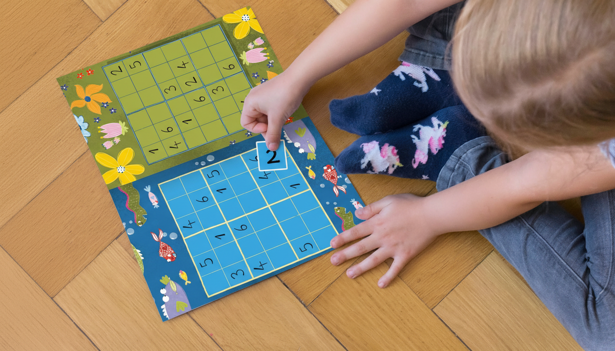 Ein Mädchen sitzt auf dem Boden. Man sieht sie von oben, wie sie in einem Stickerheft Sodoku macht.