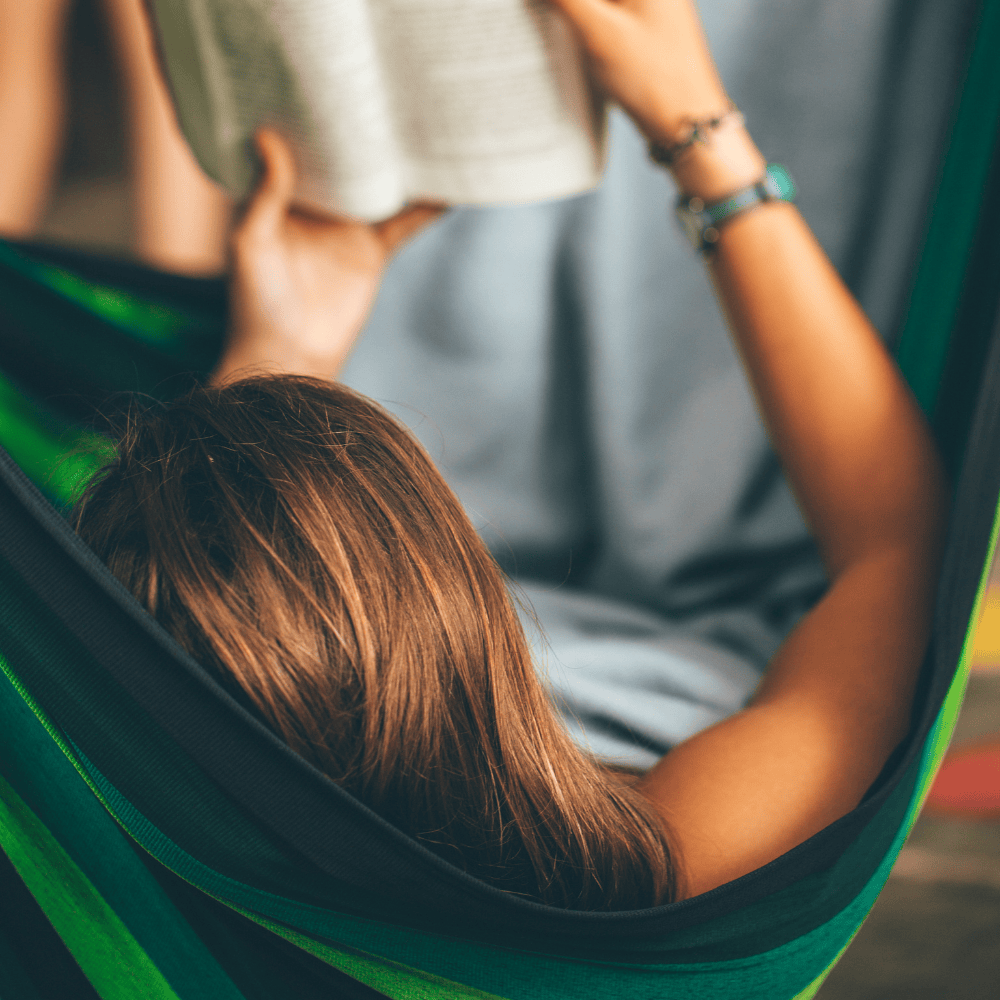 Eine Person liegt entspannt in einer grün-schwarzen Hängematte und liest ein Buch. Der Fokus liegt auf dem Rücken und den braunen Haaren der Person, während die Arme das Buch halten. Im Hintergrund ist ein unscharfer Stoff zu erkennen, der eine ruhige und entspannte Atmosphäre vermittelt.