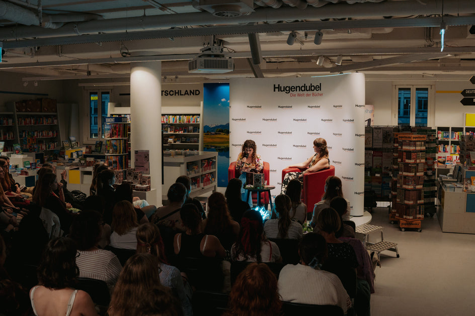 Gezeigt wird eine Lesung oder Gesprächsveranstaltung in einer Buchhandlung. Vor einem Hintergrund mit dem Schriftzug „Hugendubel – Die Welt der Bücher“ sitzen zwei Personen in roten Sesseln auf einer kleinen Bühne. Davor verfolgt ein großes Publikum die Veranstaltung, ringsum sind Bücherregale und Verkaufstische zu sehen.