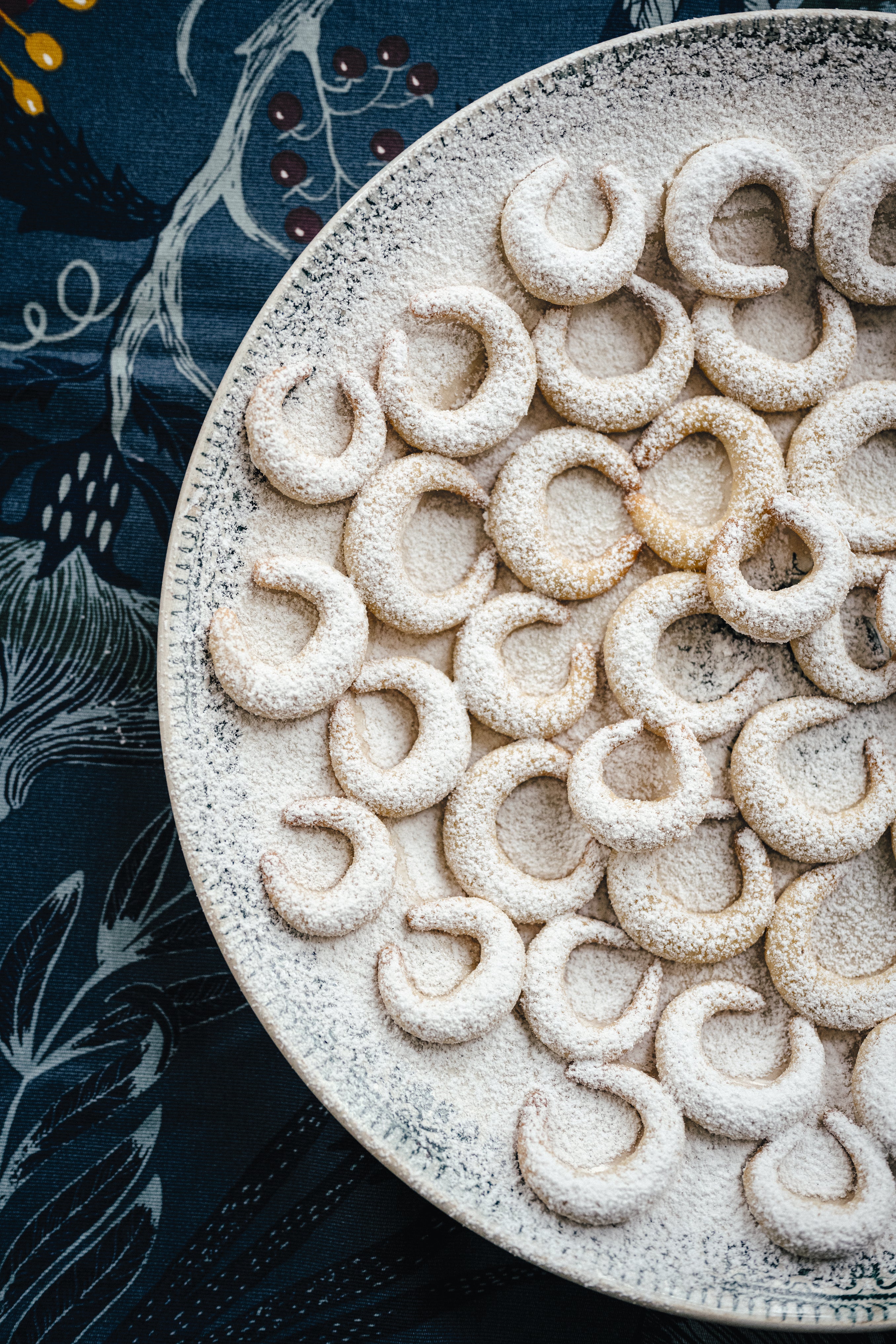 Ein großer Teller, gefüllt mit Vanillekipferln, die mit Puderzucker bestäubt sind. Die halbmondförmigen Kekse liegen dicht beieinander, während der Teller auf einem dunklen, gemusterten Stoff mit floralen und beerenartigen Motiven arrangiert ist, was eine elegante und festliche Atmosphäre schafft.