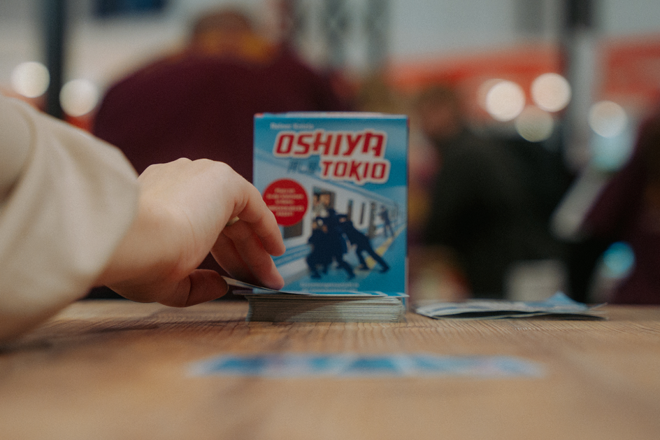Im Vordergrund ist eine Hand zu sehen, die eine Karte von einem Stapel nimmt. Im Hintergrund ist das zugehörige Spiel. Zu lesen ist Oshiya Tokio auf dem Karton.
