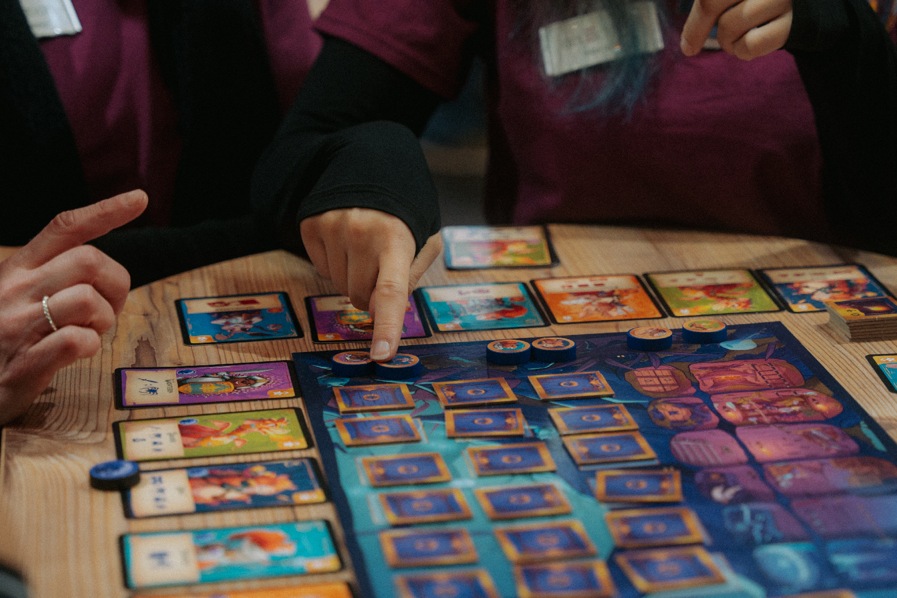 Ein ausgebreitetes Spiel auf einem Tisch. Zu sehen sind Karten, ein Spielbrett und Spielfiguren. Im Anschnitt sieht man zwei Menschen, die gerade spielen.