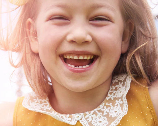 Ein Kind trägt ein gelbes Kleid mit weißen Punkten und einem dekorativen Spitzenkragen. Die Szene ist in warmes Licht getaucht, das eine sanfte, sommerliche Atmosphäre vermittelt.