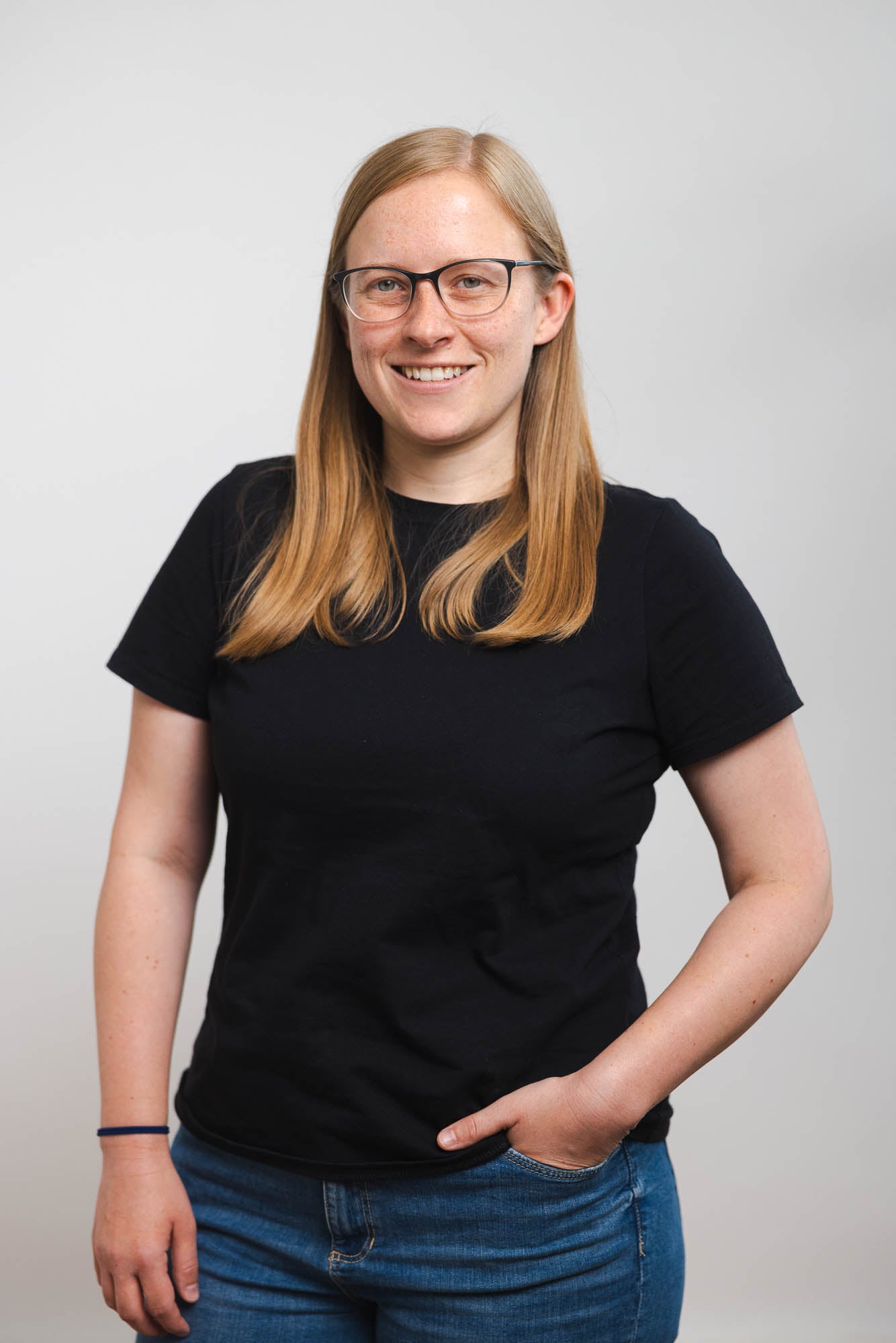 Eine Frau mit hellblondem, glattem, schulterlangem Haar trägt ein schwarzes T-Shirt und eine blaue Jeans. Ihre linke Hand steckt lässig in der Hosentasche. Sie steht vor einem neutralen, grauen Hintergrund.