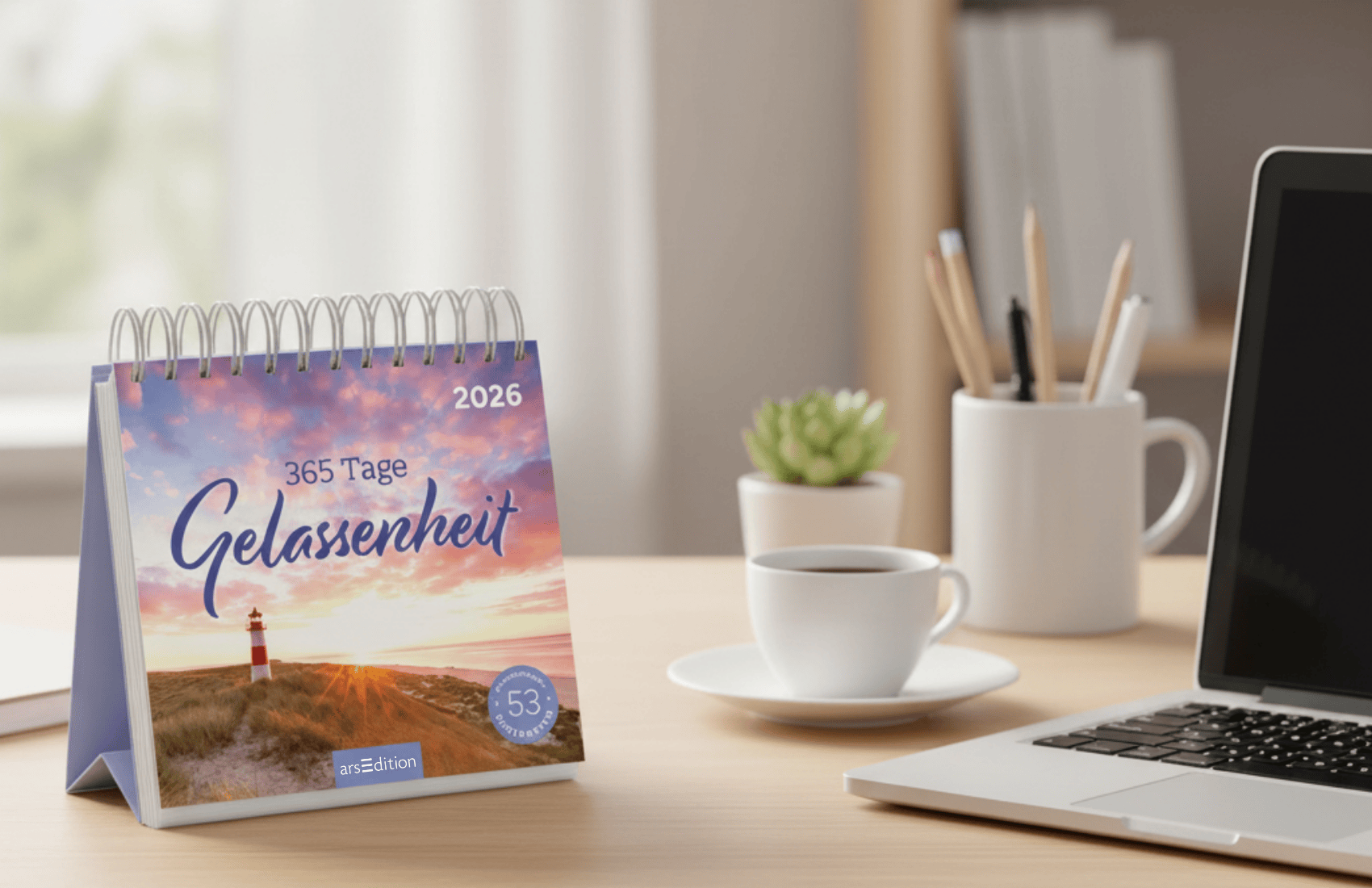 Ein Schreibtisch aus hellem Holz. Links im Bild der Postkartenkalender 365 Tage Gelassenheit, rechts ein Laptop, im Hintergrund eine Tasse, eine Pflanze und ein Stiftebecher.