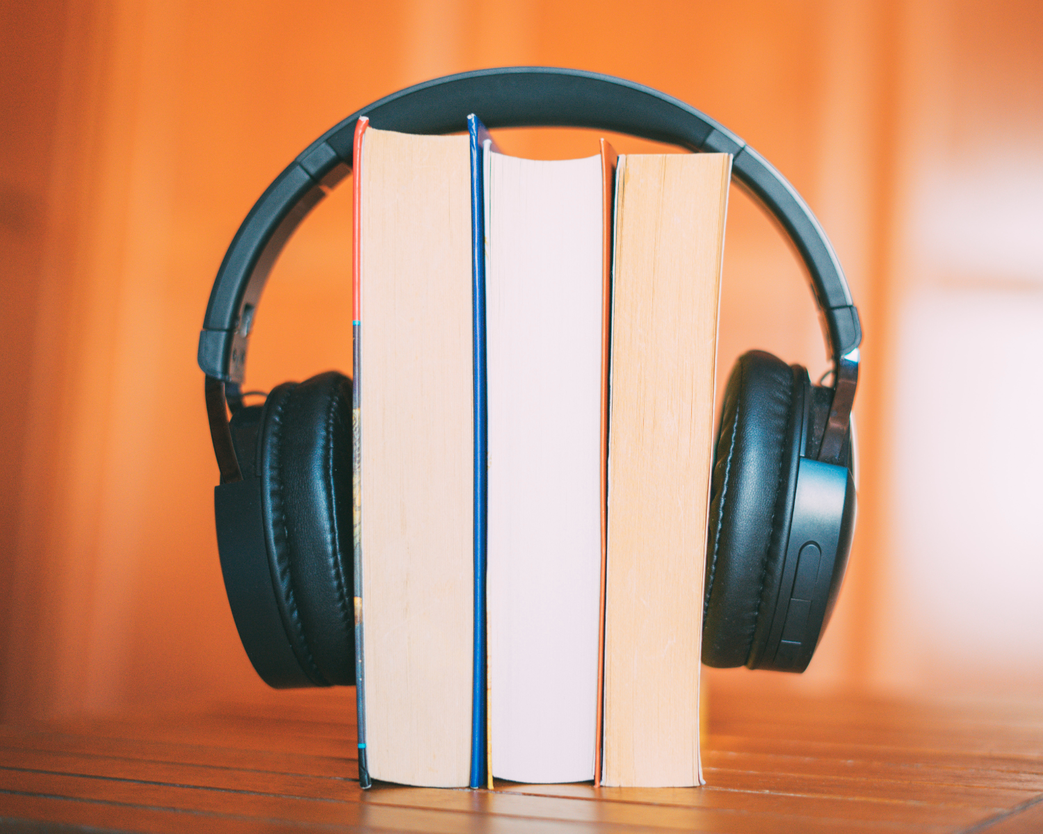 Ein Paar schwarze Over-Ear-Kopfhörer, die aufrecht an drei nebeneinander stehenden Büchern lehnen. Die Szene ist vor einem warmen, orangefarbenen Hintergrund aufgenommen.