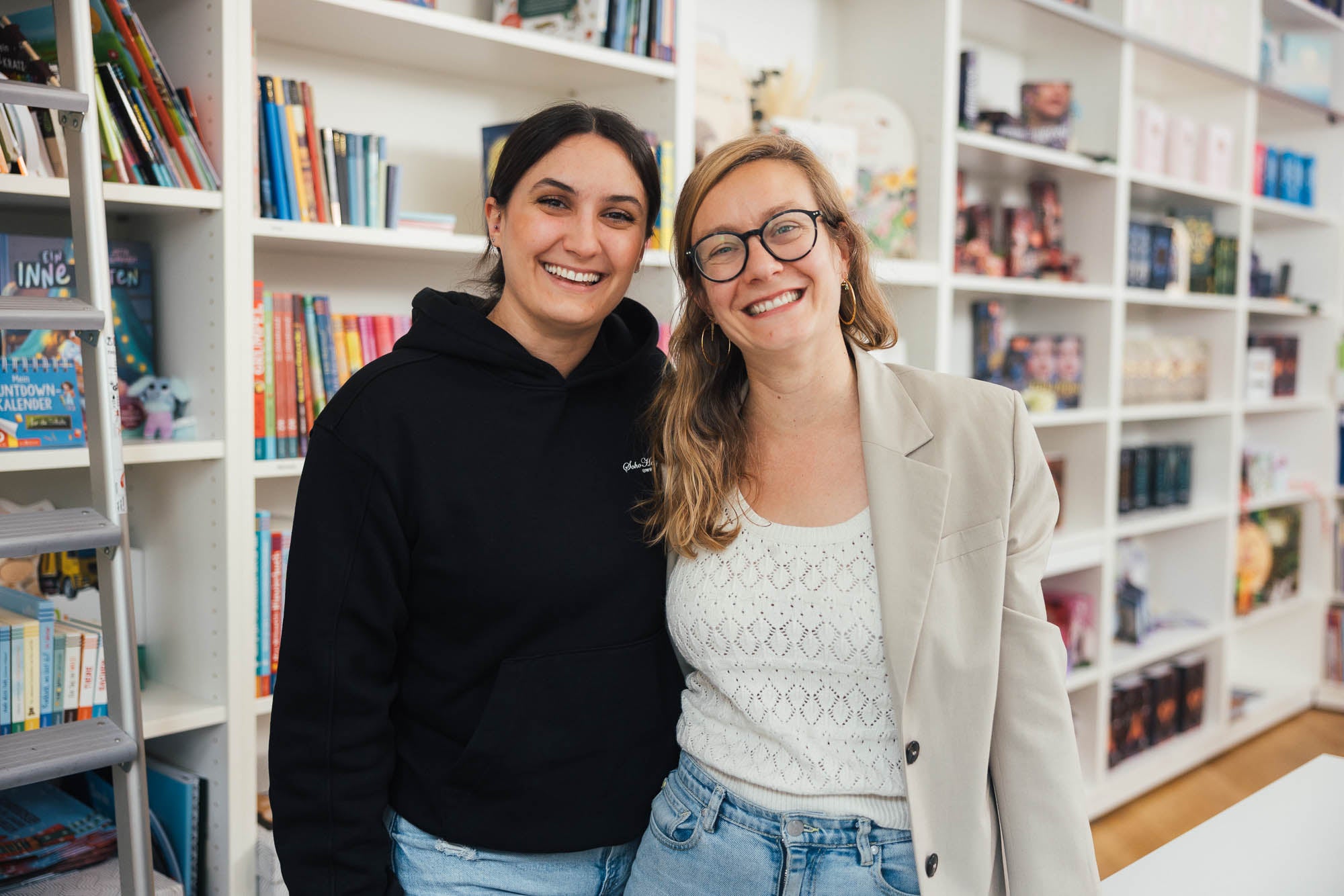 Zwei Frauen stehen nebeneinander vor einer Bücherregalwand. Sie lächeln in die Kamera. Die Regale im Hintergrund sind mit bunten Büchern gefüllt und schaffen eine freundliche und offene Umgebung.
