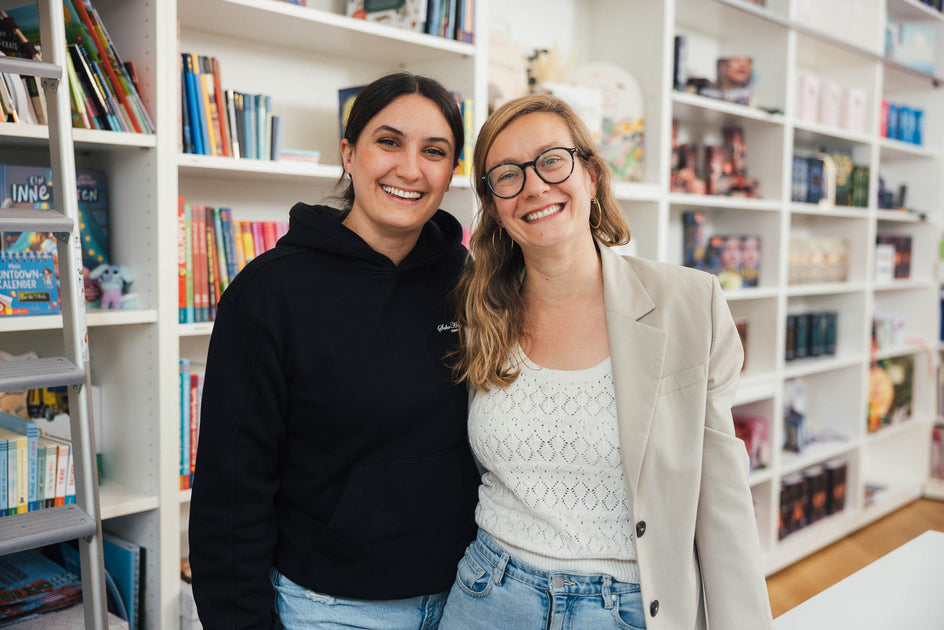 Zwei Frauen stehen nebeneinander vor einer Bücherregalwand. Sie lächeln in die Kamera. Die Regale im Hintergrund sind mit bunten Büchern gefüllt und schaffen eine freundliche und offene Umgebung.