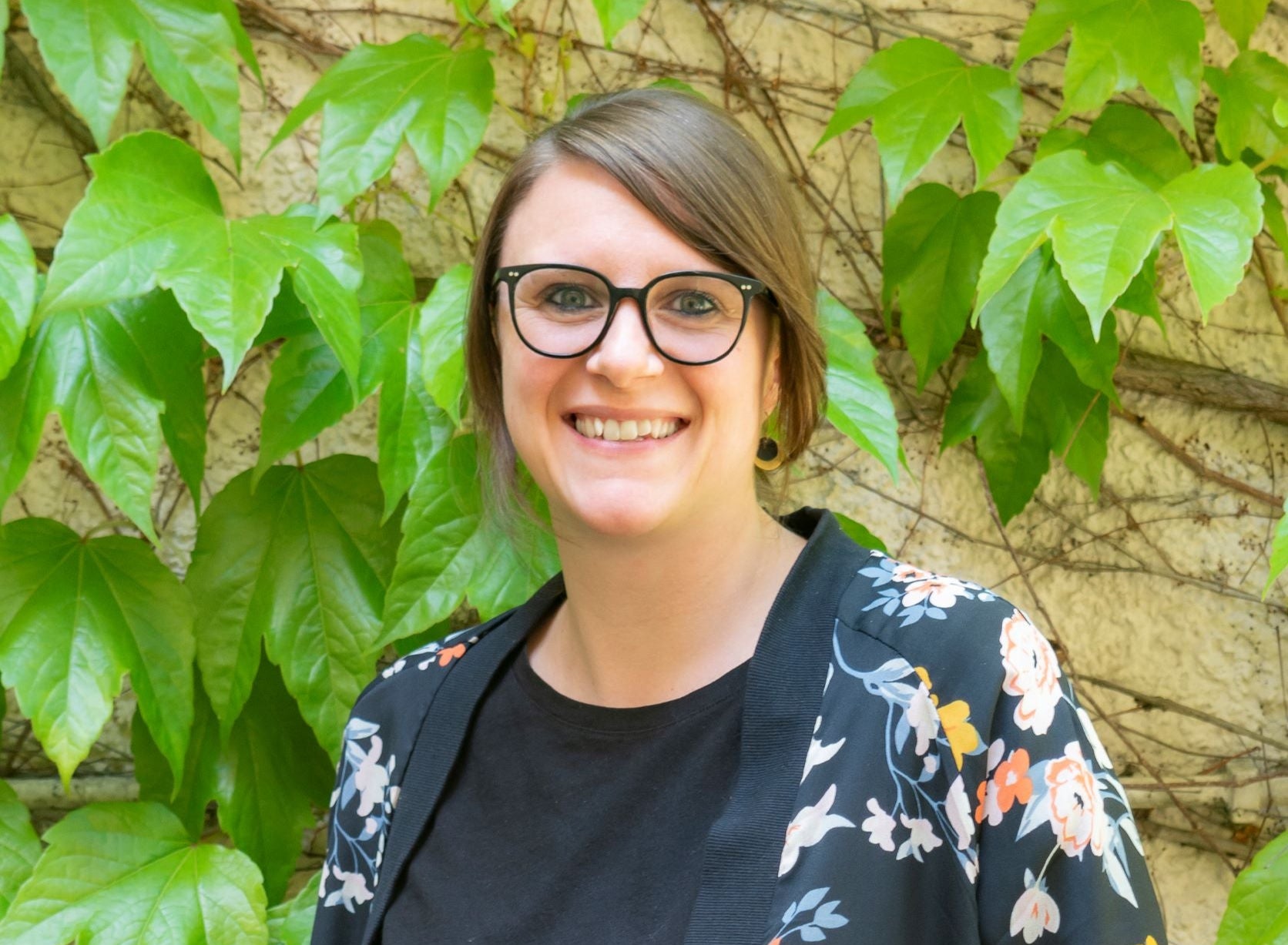 Sandra Kauer steht vor einer mit grünem Efeu bewachsenen Wand. Sie trägt ein schwarzes Oberteil und eine Jacke mit floralem Muster.
