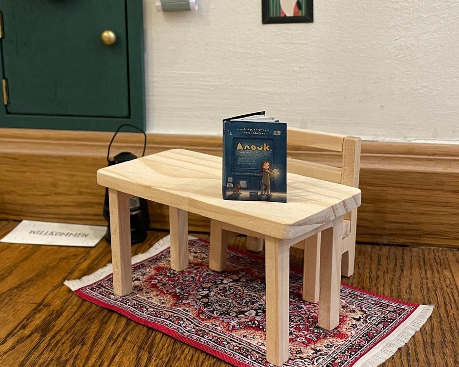 Miniaturszene mit einem kleinen Holztisch und einer passenden Holzbank, die auf einem detailreichen, orientalischen Teppich steht. Auf dem Tisch liegt ein aufgeschlagenes Buch mit dem Titel 'Anouk'. Im Hintergrund sind eine grüne Tür mit goldfarbenem Griff, ein Bilderrahmen und ein schwarzes Objekt zu sehen. Die Szene ist liebevoll gestaltet und wirkt wie eine winzige Wohnumgebung."