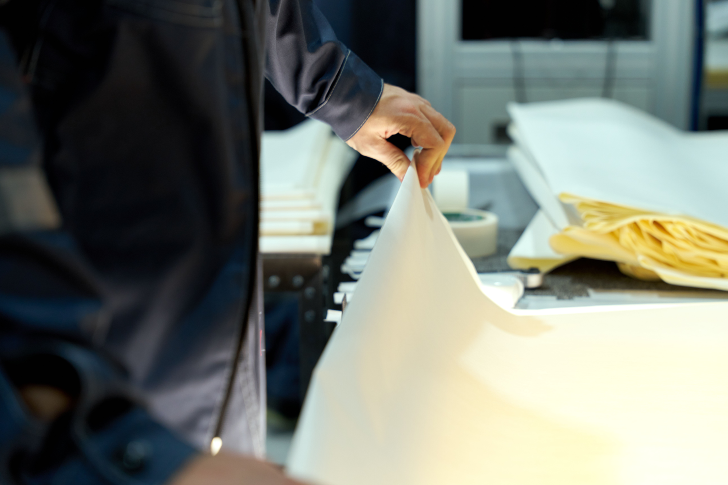 Eine Person in dunkler Arbeitskleidung greift in einer Produktionsumgebung nach einem großen, hellen Papierbogen. Im Hintergrund liegen weitere gestapelte Bögen auf einer Maschine oder Arbeitsfläche, die Szene ist nah aufgenommen und zeigt vor allem Hand, Material und Arbeitsprozess.