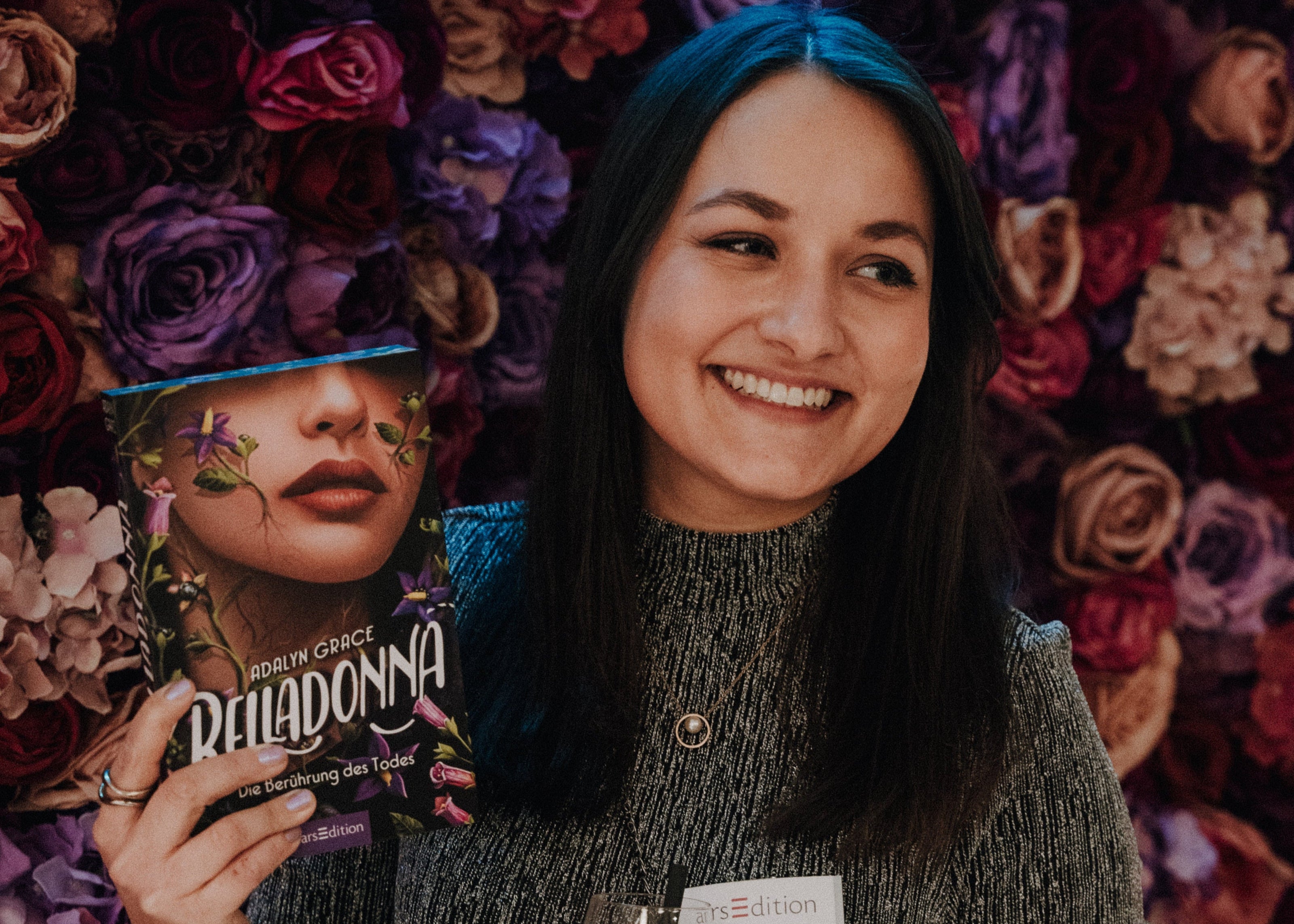 Eine Person hält das Buch „Belladonna – Die Berührung des Todes“ von Adalyn Grace in der Hand. Im Hintergrund ist eine dekorative Wand aus verschiedenen Blumen in Rosa-, Rot- und Lilatönen zu sehen. Die Person trägt ein graues, strukturiertes Oberteil und ein Namensschild der Marke „arsEdition“. Der Fokus liegt auf dem Buch und der blumigen Kulisse.