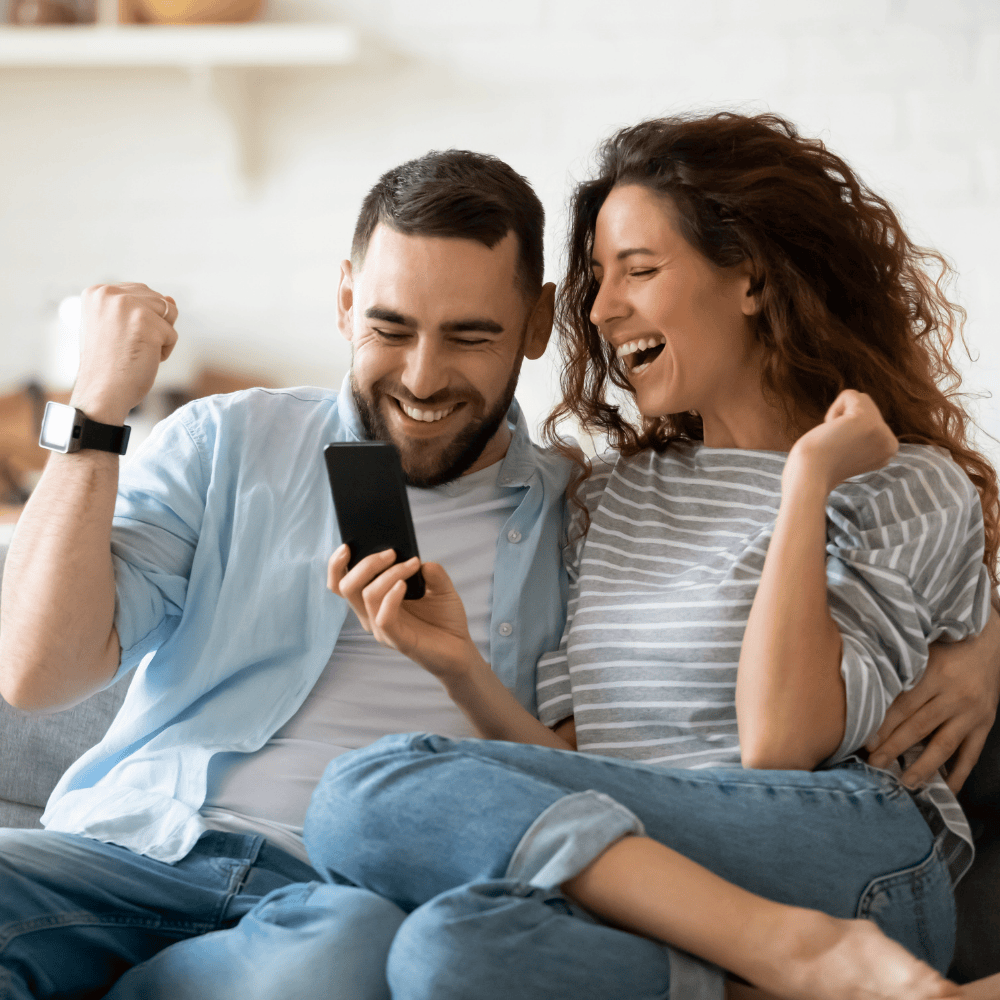 Ein Paar sitzt auf einem Sofa und feiert gemeinsam einen Erfolg. Beide halten ein Smartphone in der Hand, während sie jubeln und die Faust triumphierend ballen. Die Szene vermittelt Freude und eine positive Stimmung in einem gemütlichen Wohnumfeld.