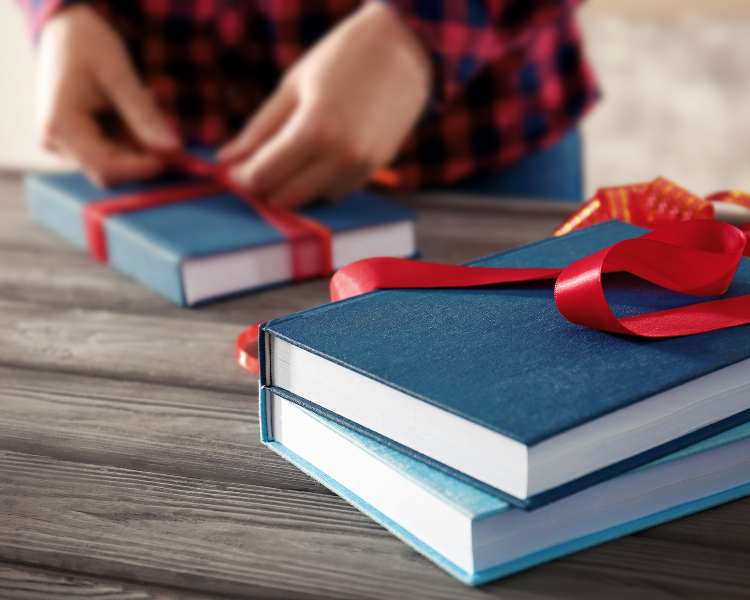 Zwei blaue Bücher mit weißem Buchschnitt, die auf einem Holztisch liegen. Ein rotes Geschenkband liegt lose um eines der Bücher, während im Hintergrund unscharf Hände ein weiteres Buch mit rotem Band verpacken.