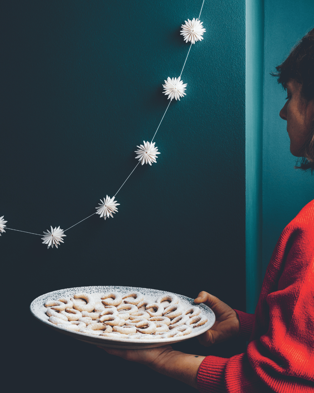 Eine Frau in einem roten Pullover trägt einen Teller mit Vanillekipferln, die mit Puderzucker bestäubt sind