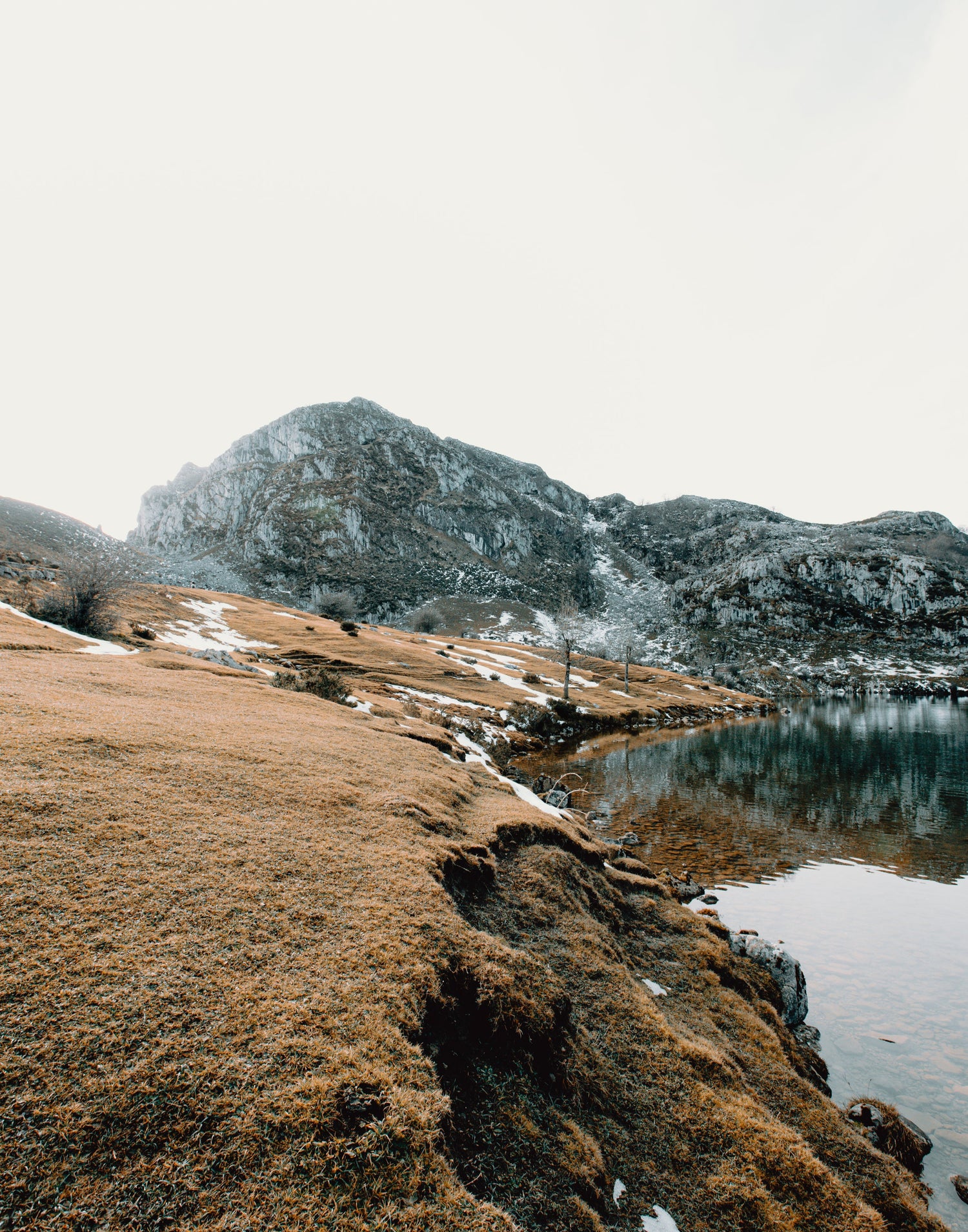 Landschaftsaufnahme eines ruhigen Bergsees mit klarer Spiegelung, umgeben von schneebedeckten Hügeln und felsigen Bergen im Hintergrund. Der Himmel ist hell und leicht bewölkt.