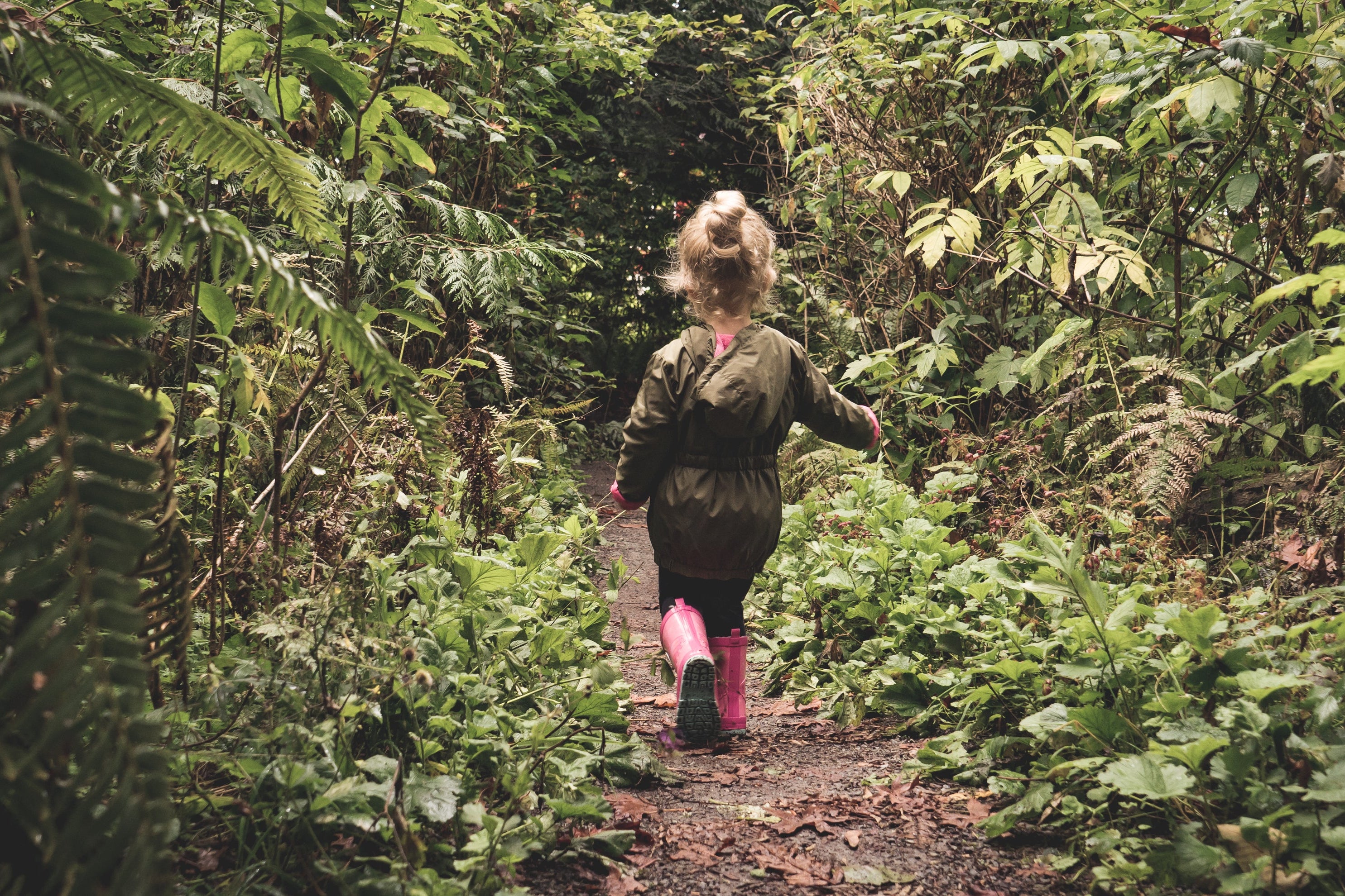 Ein kleines Kind mit blonden Haaren, das zu einem Zopf gebunden ist, läuft auf einem schmalen, von grünen Pflanzen und Bäumen umgebenen Waldweg. Das Kind trägt eine olivgrüne Jacke und pinke Gummistiefel. Die Szene vermittelt eine ruhige, naturverbundene Atmosphäre.