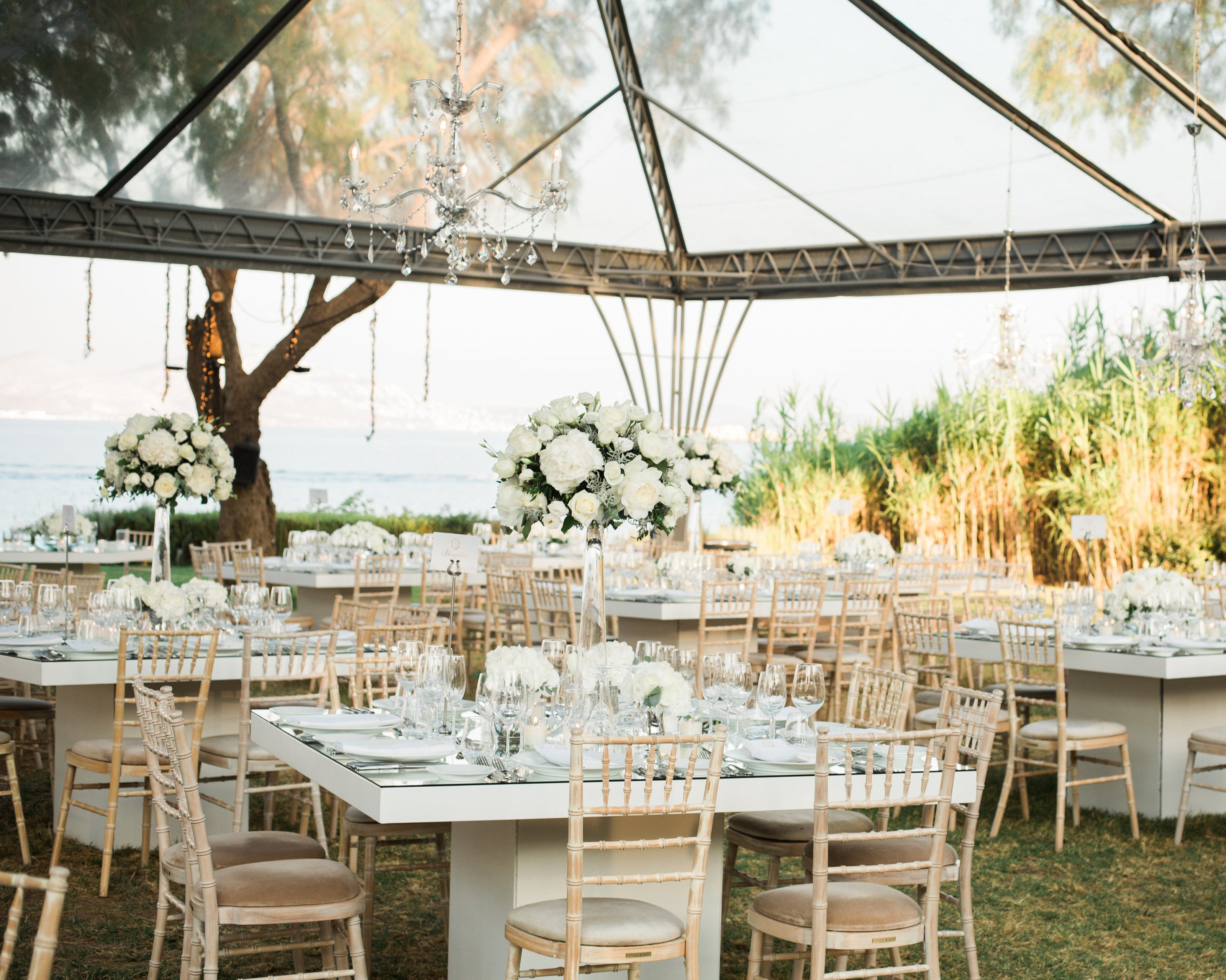 An elegant outdoor event setup features white tables adorned with floral centerpieces of white roses and greenery. The tables are surrounded by bamboo-style chairs, and crystal chandeliers hang under a transparent canopy. In the background, there are trees, tall grass, and a view of the sea.