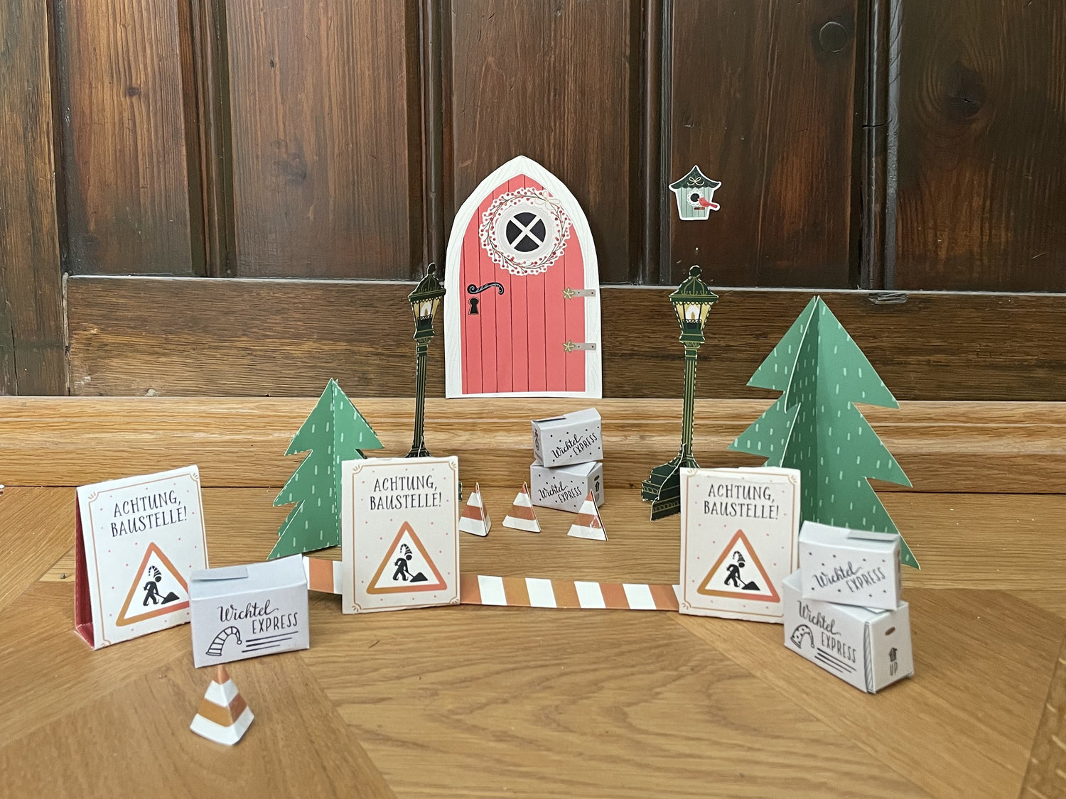 Vor einer roten, bogenförmigen Wichteltür an einer dunklen Holzwand steht eine kleine, weihnachtliche Baustellen-Szene aus Papier: zwei grüne Tannen, zwei Laternen, mehrere Warnschilder mit der Aufschrift „Achtung, Baustelle!“, ein rot-weißes Absperrband und mehrere orange-weiße Pylonen. Daneben liegen kleine Pakete/Kartons mit der Aufschrift „Wichtel Express“ auf dem Holzboden.