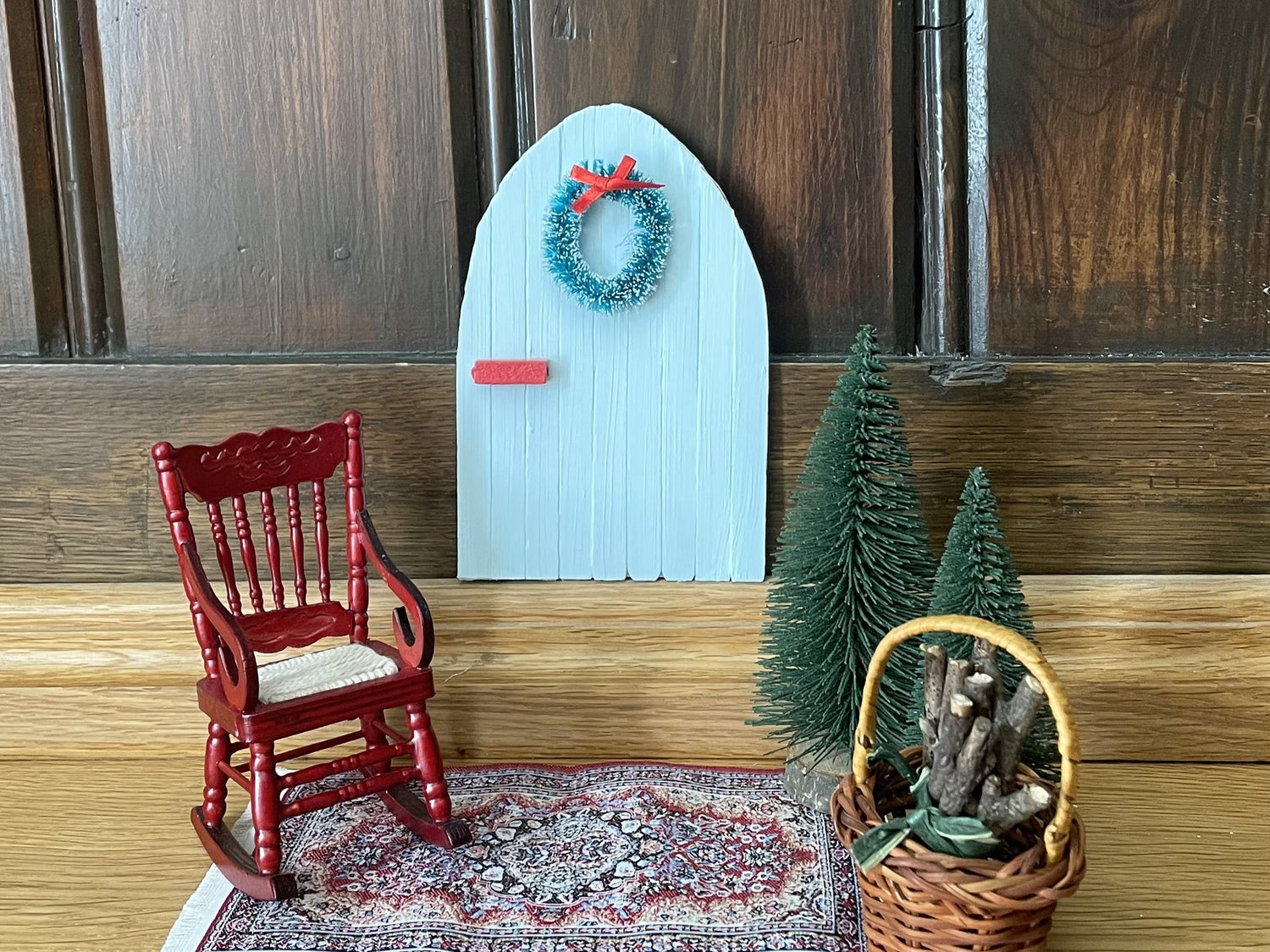 Vor einer dunkelbraunen Holzwand steht eine kleine Tür in Hellblau mit einem kleinen Kranz und einer roten Türklinke. Daneben stehen ein kleiner roter Stuhl und Miniatur-Weihnachtsbäume. Die Szene ist eine Wichteltür.