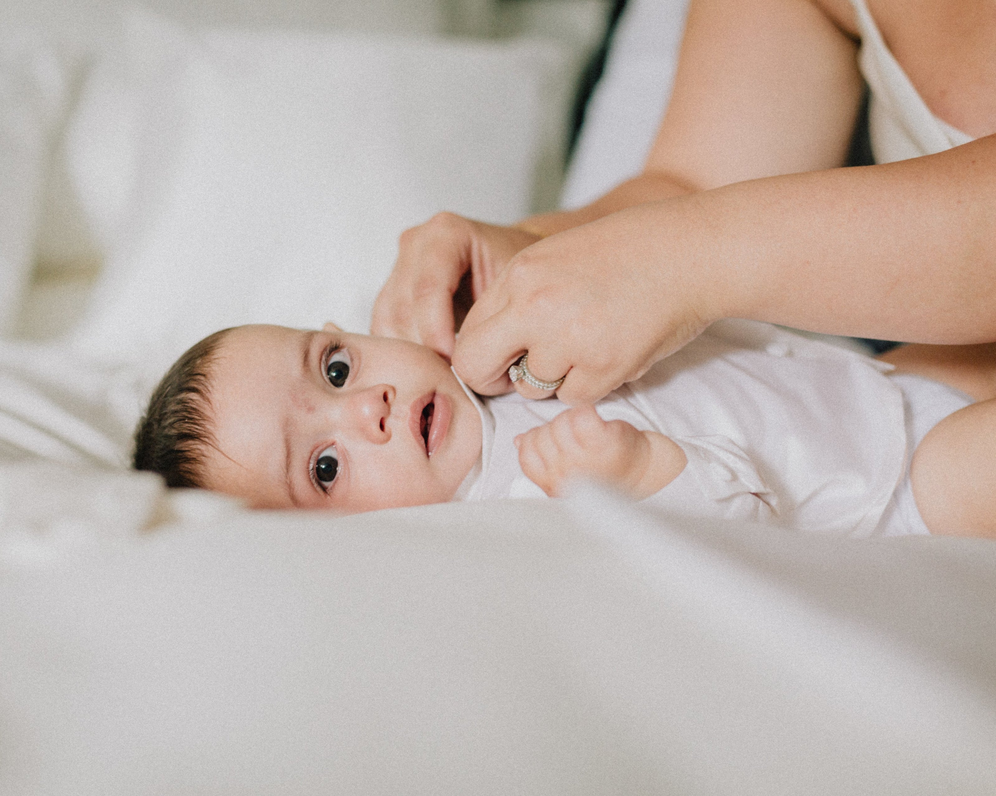 A close-up image shows an adult's hands gently dressing a baby in a white onesie. The baby lies on a soft white surface, and the adult is wearing a ring on one hand, adding a touch of detail to the scene.
