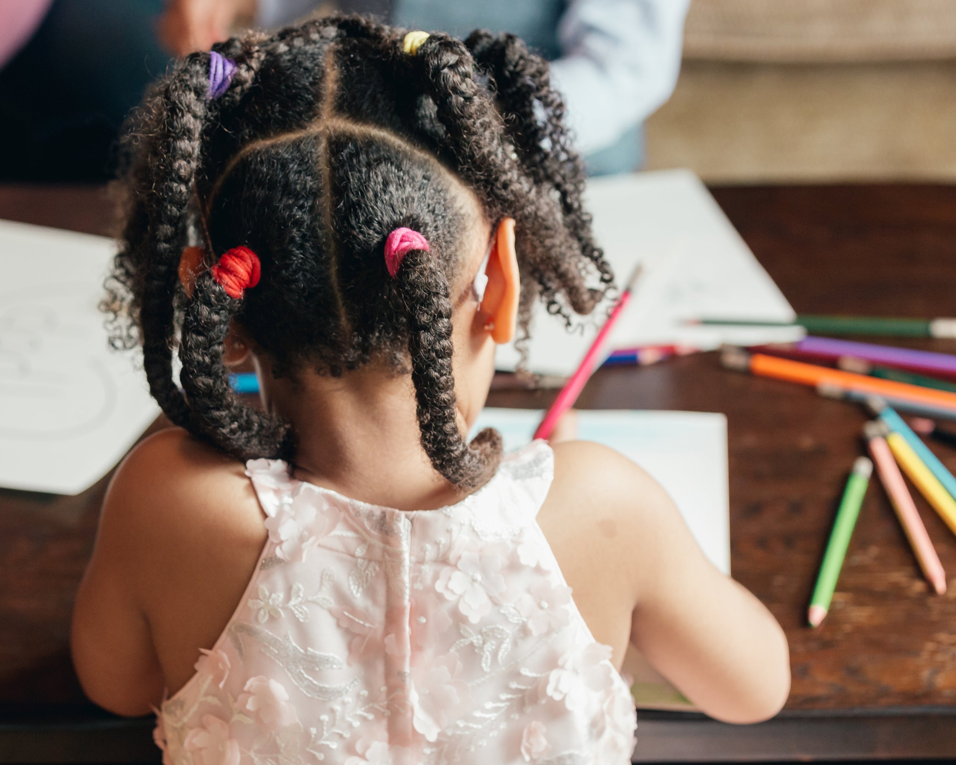 Ein Kind sitzt an einem Tisch und malt mit bunten Buntstiften auf Papier. Es trägt ein ärmelloses, blassrosa Kleid mit Blumenmuster. Die Frisur besteht aus mehreren Zöpfen, die mit bunten Haargummis zusammengebunden sind. Im Hintergrund liegen verstreut weitere Stifte und Papier auf dem Tisch.