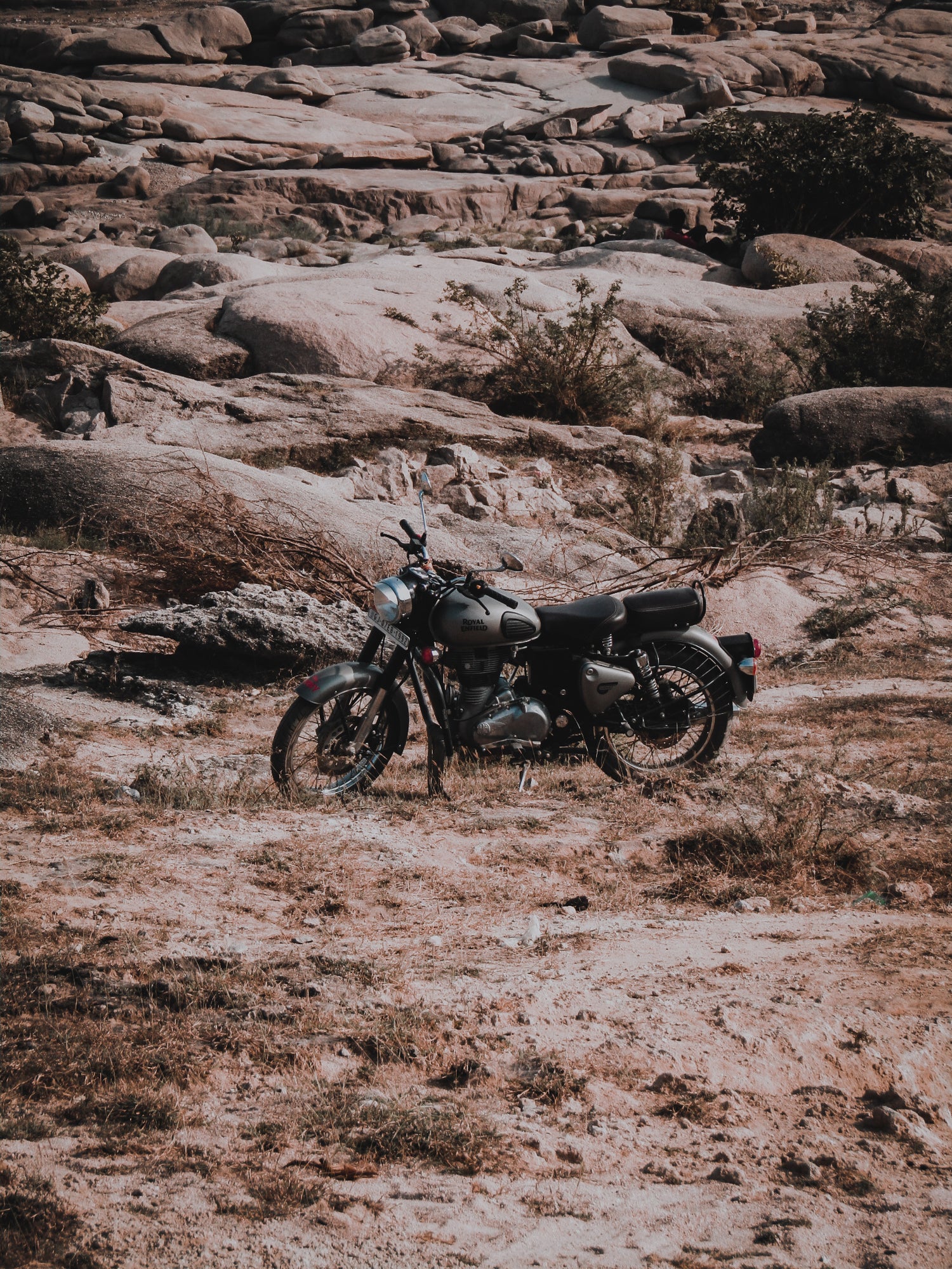 Ein klassisches Motorrad in mattem Grau steht auf einer sandigen, trockenen Fläche mit Felsen und spärlicher Vegetation im Hintergrund.