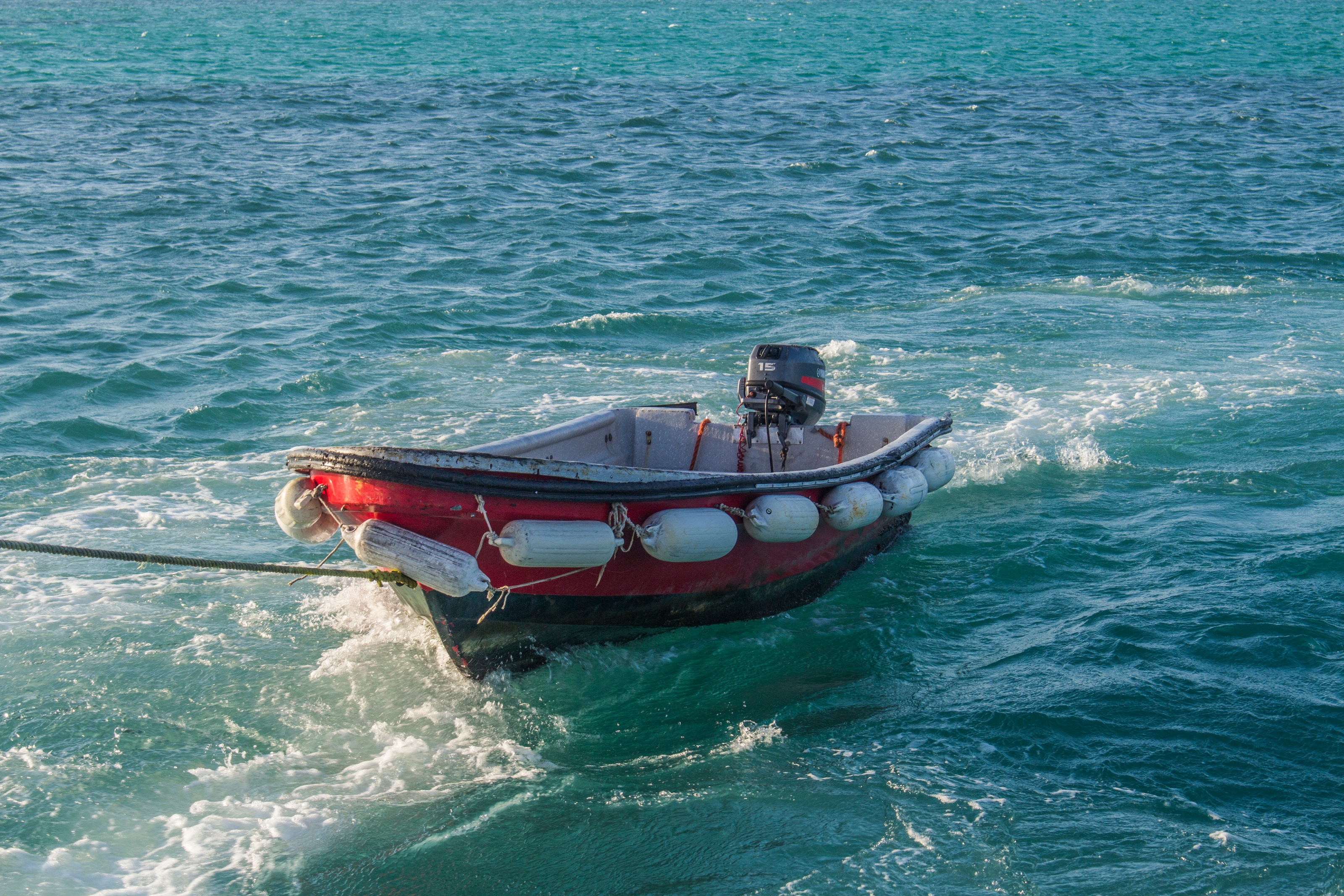 Ein kleines, rotes Motorboot mit weißen Fendern treibt auf türkisblauem, leicht welligem Wasser. Am Heck des Bootes ist ein Außenbordmotor befestigt. Das Boot ist mit einem Seil an etwas außerhalb des Bildes festgebunden.
