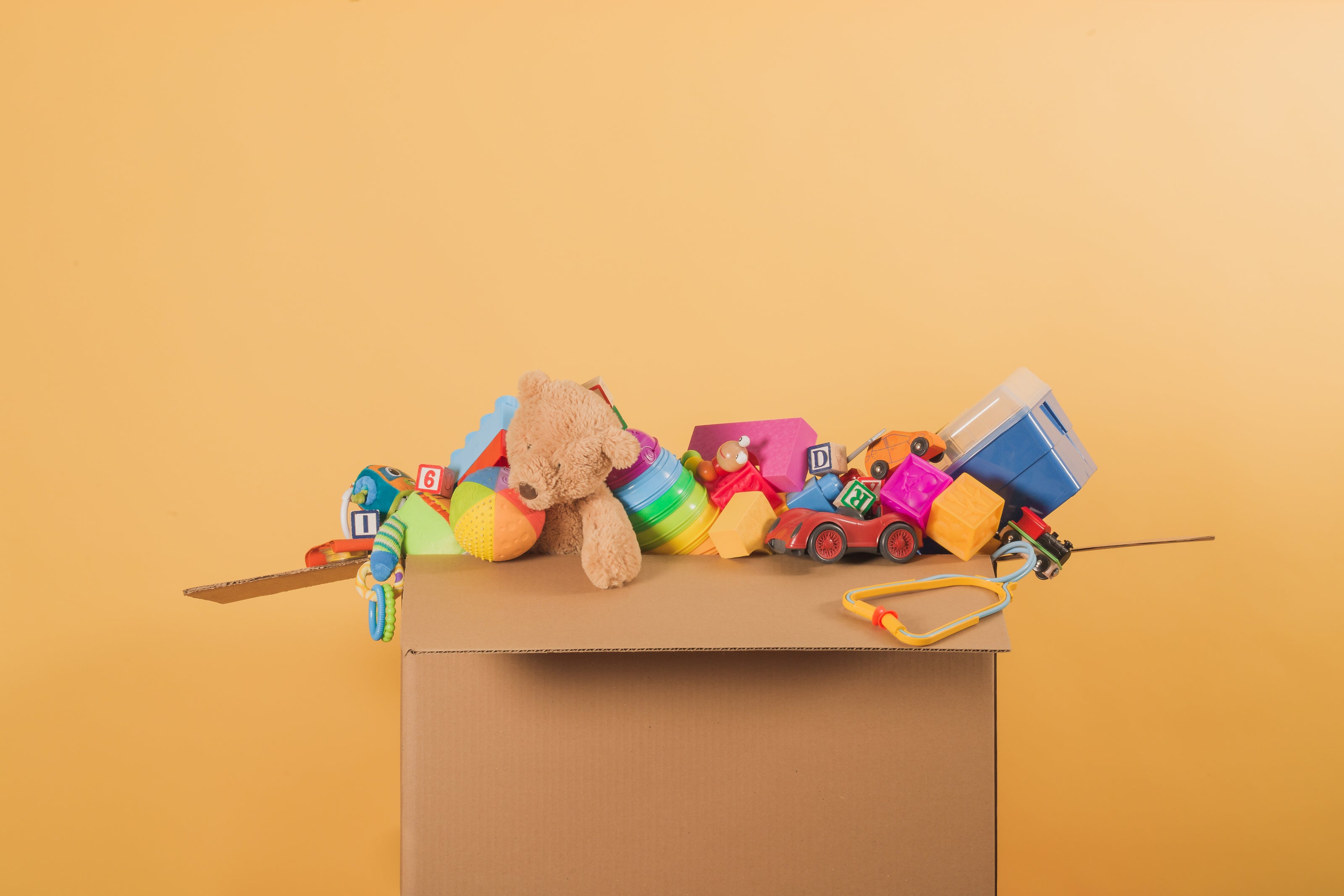 Ein geöffneter Karton gefüllt mit verschiedenen bunten Kinderspielzeugen. Zu sehen sind unter anderem ein Teddybär, ein Regenbogen-Stapelspiel, ein rotes Spielzeugauto, Bauklötze und ein Spielzeug-Stethoskop. Der Hintergrund ist in einem warmen Gelbton gehalten