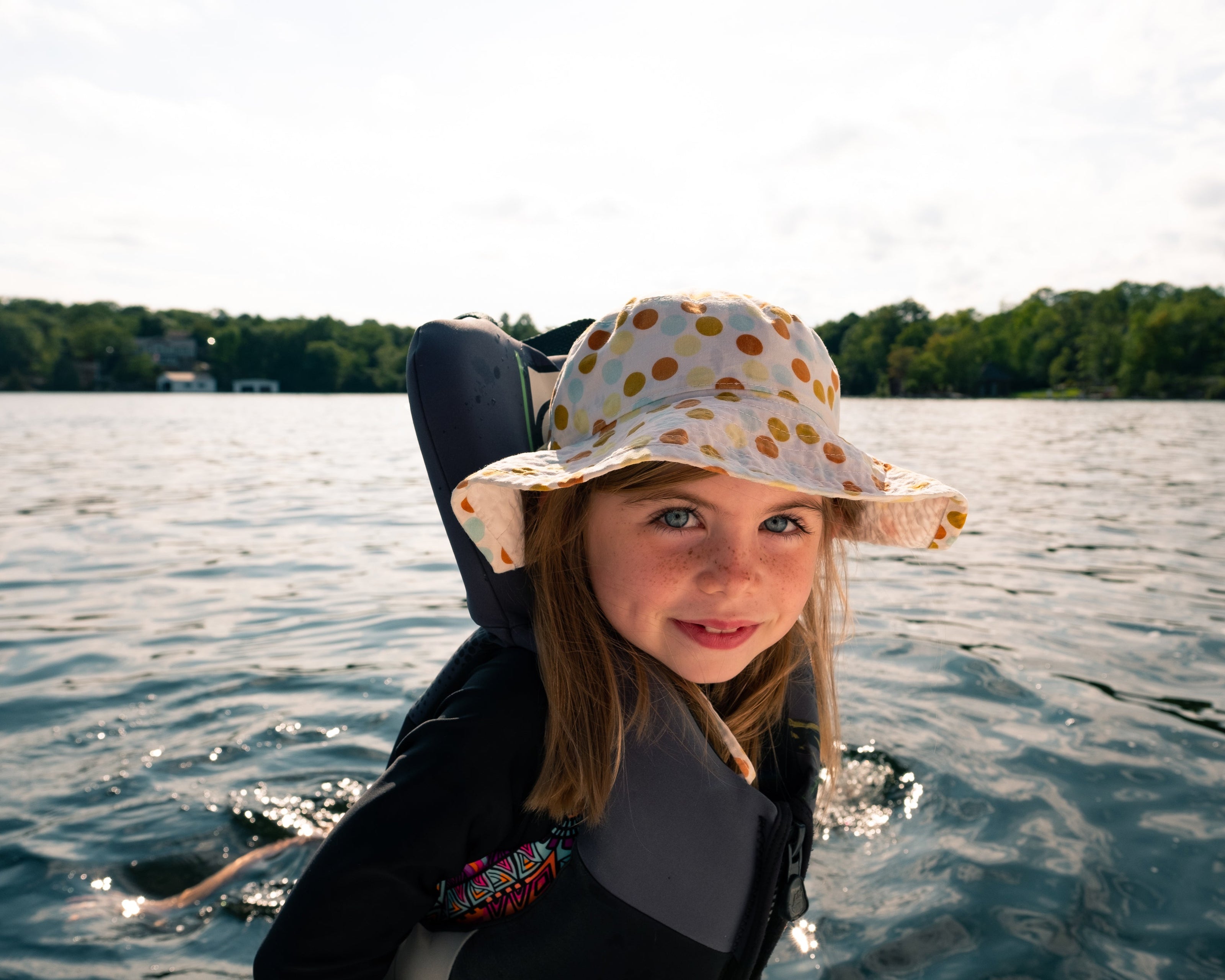 Ein Kind trägt eine gepunktete Sonnenmütze und eine Schwimmweste, während es sich auf einem Gewässer befindet. Der Hintergrund zeigt eine ruhige, bewaldete Uferlandschaft und ein weitläufiges Gewässer unter einem hellen Himmel. Die Szene vermittelt einen sommerlichen und entspannten Moment auf dem Wasser.