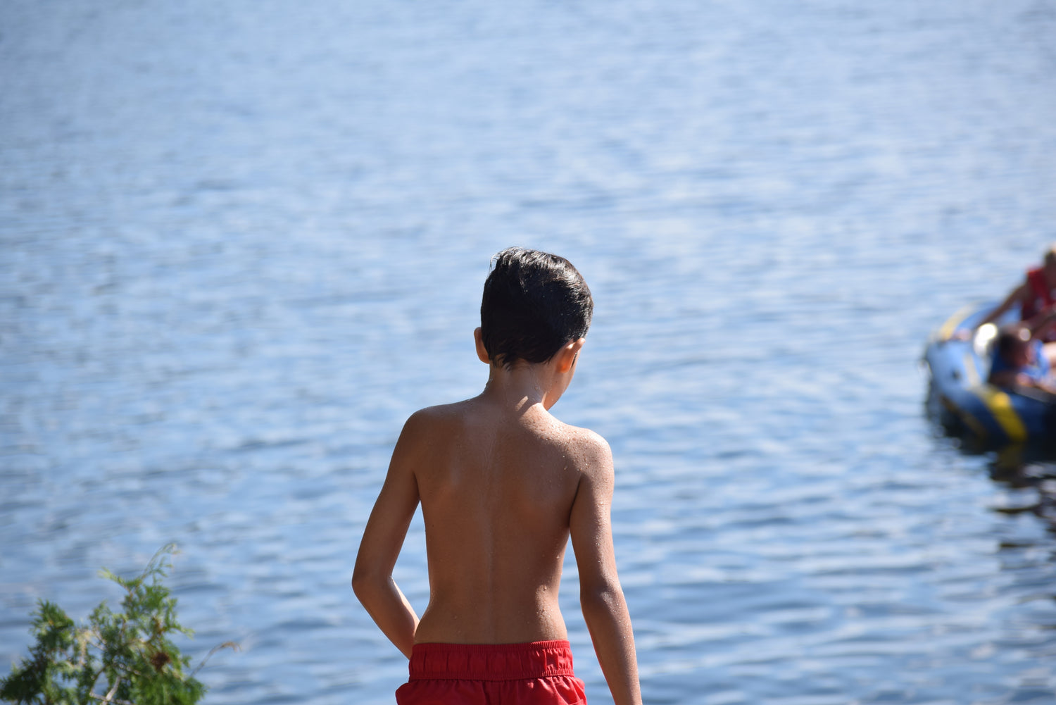 Ein Junge mit kurzen dunklen Haaren steht mit dem Rücken zur Kamera am Ufer eines Sees. Er trägt eine rote Badehose und blickt auf das glitzernde Wasser.