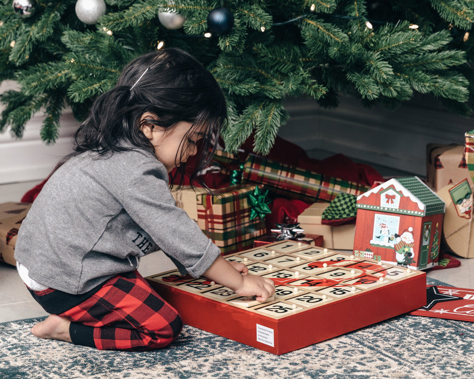 Ein Kind sitzt auf einem gemusterten Teppich vor einem geschmückten Weihnachtsbaum und spielt mit einem hölzernen Adventskalender. Um den Baum herum liegen verpackte Geschenke mit festlichen Designs.