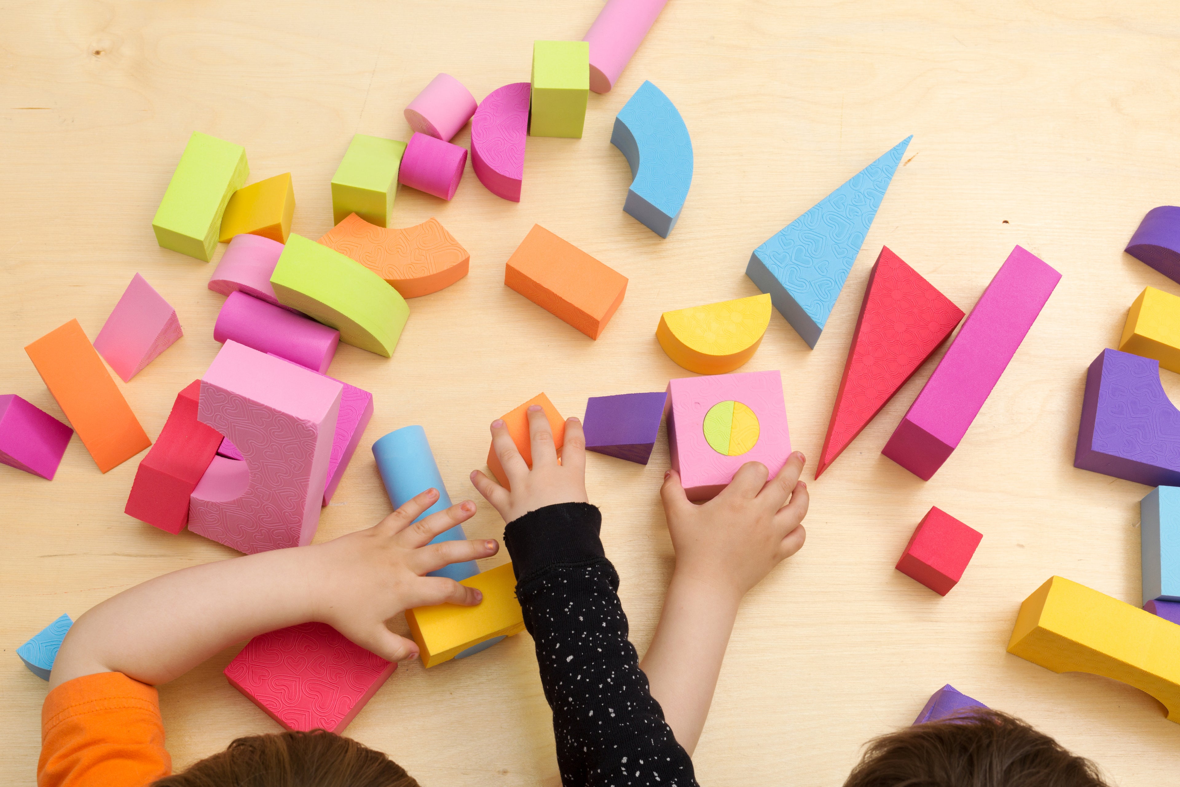 Drei Kinderhände greifen nach bunten Schaumstoffbausteinen in verschiedenen Formen und Farben, die auf einem hellen Holztisch verstreut liegen. Die Bausteine sind in leuchtendem Orange, Gelb, Pink, Blau, Grün und Lila gehalten und haben Formen wie Würfel, Zylinder, Dreiecke und Halbkreise.