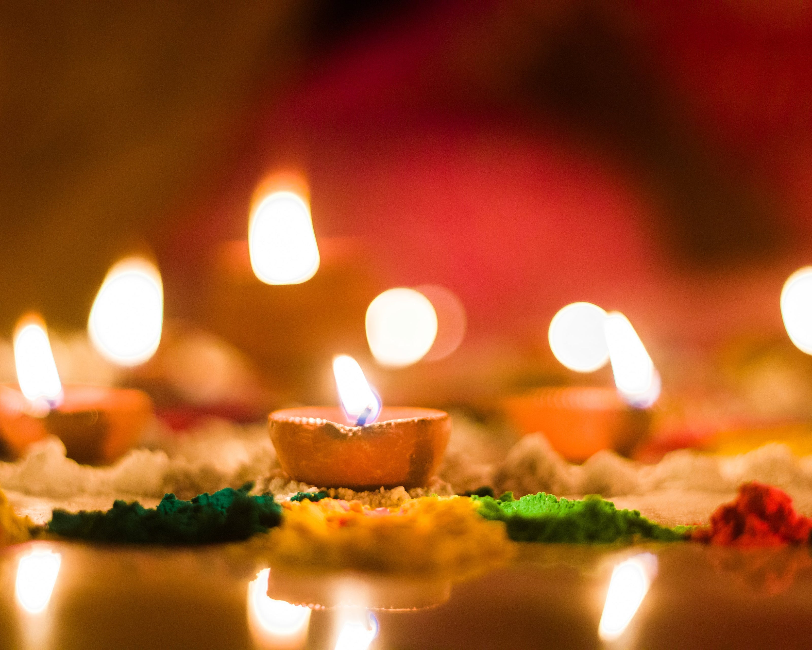 A close-up image of traditional clay oil lamps (diyas) lit with bright flames. The lamps are surrounded by vibrant colored powders, creating an artistic and festive atmosphere. The background has a warm, blurred glow.